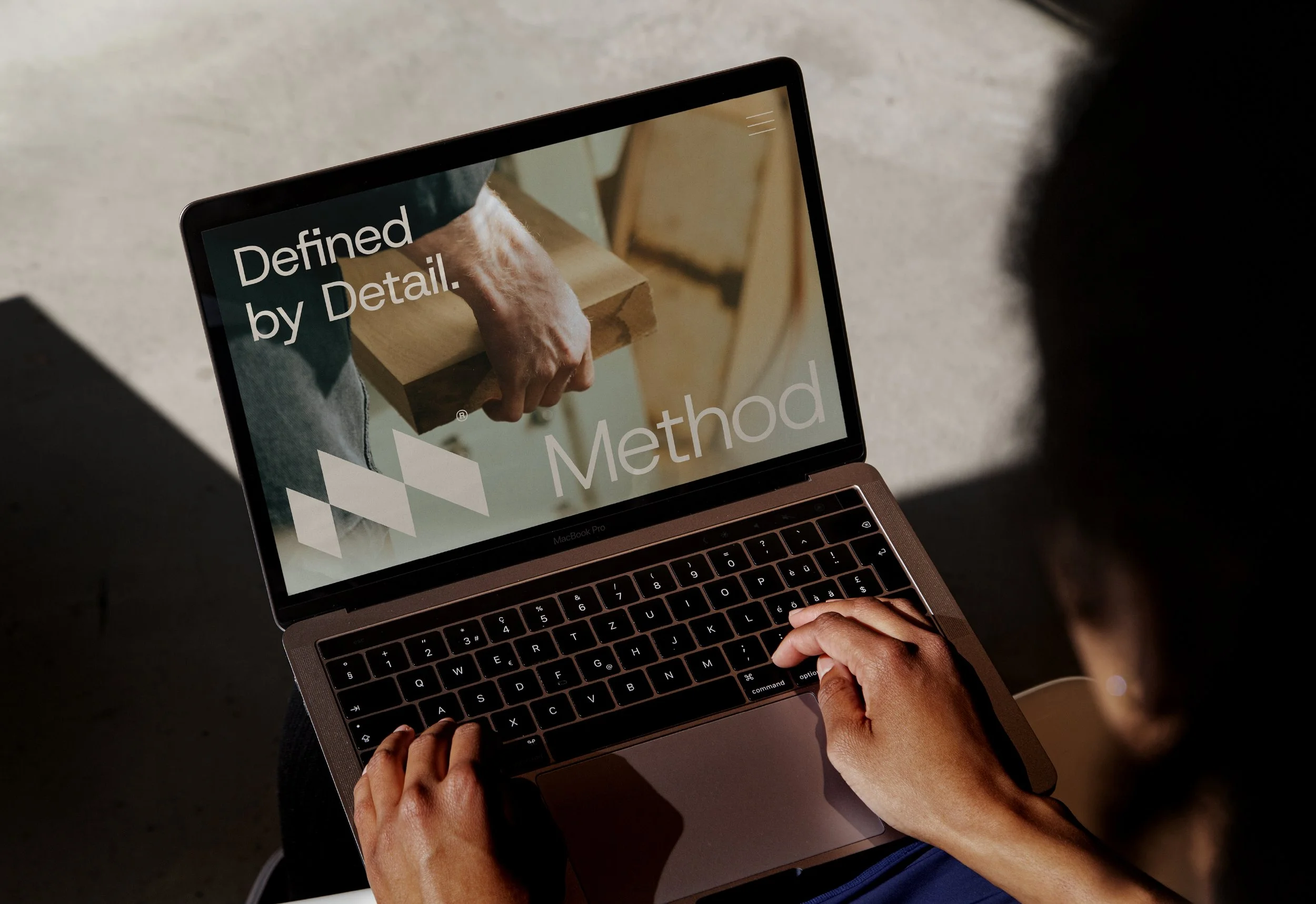 Close-up of a person using a laptop, with the screen displaying text 'Defined by Detail' and part of the word 'Method'. The person's hands are on the keyboard.