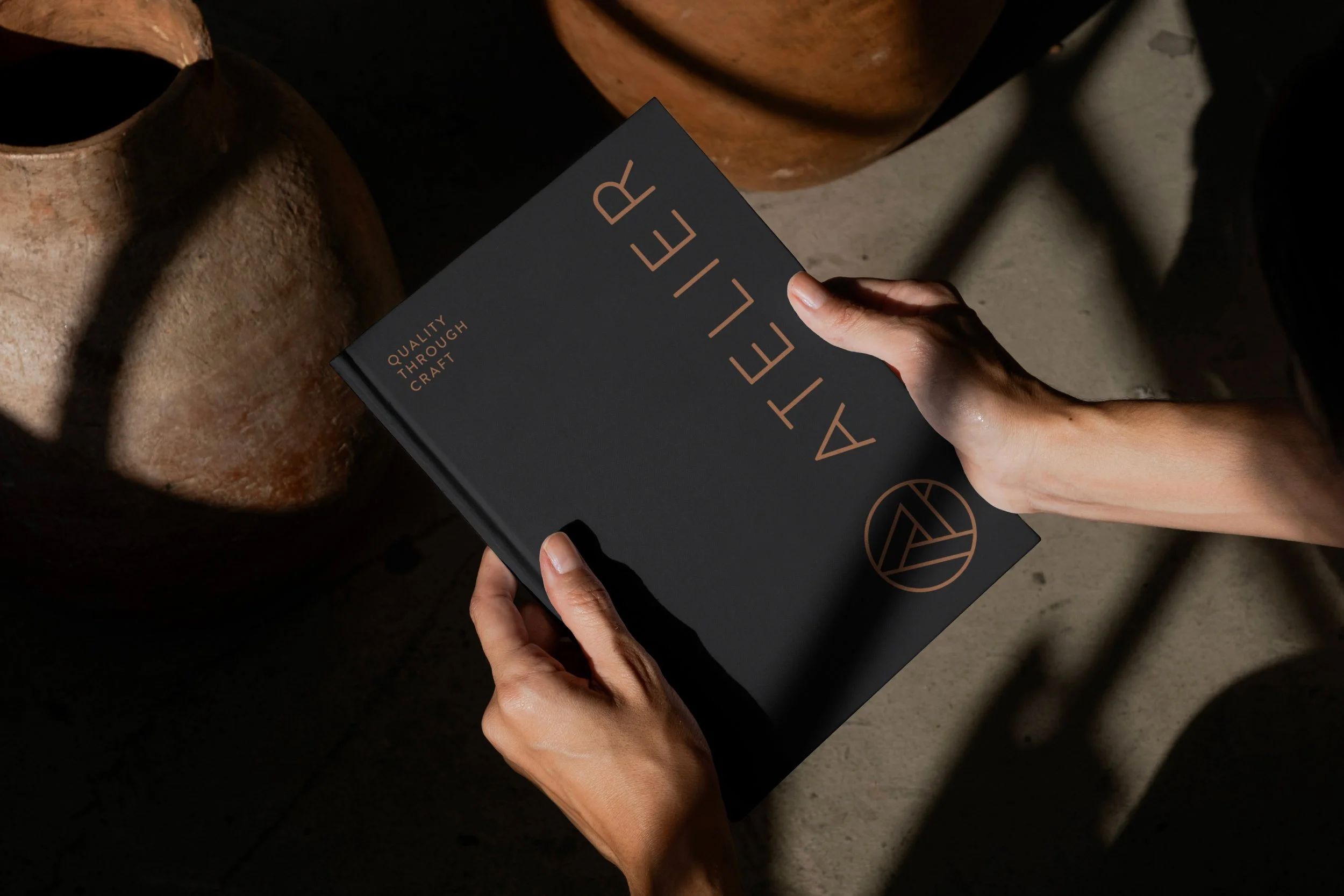 Person holding a black booklet titled 'Artelier' with the subtitle 'Quality Through Craft', surrounded by large clay pots on a concrete floor.