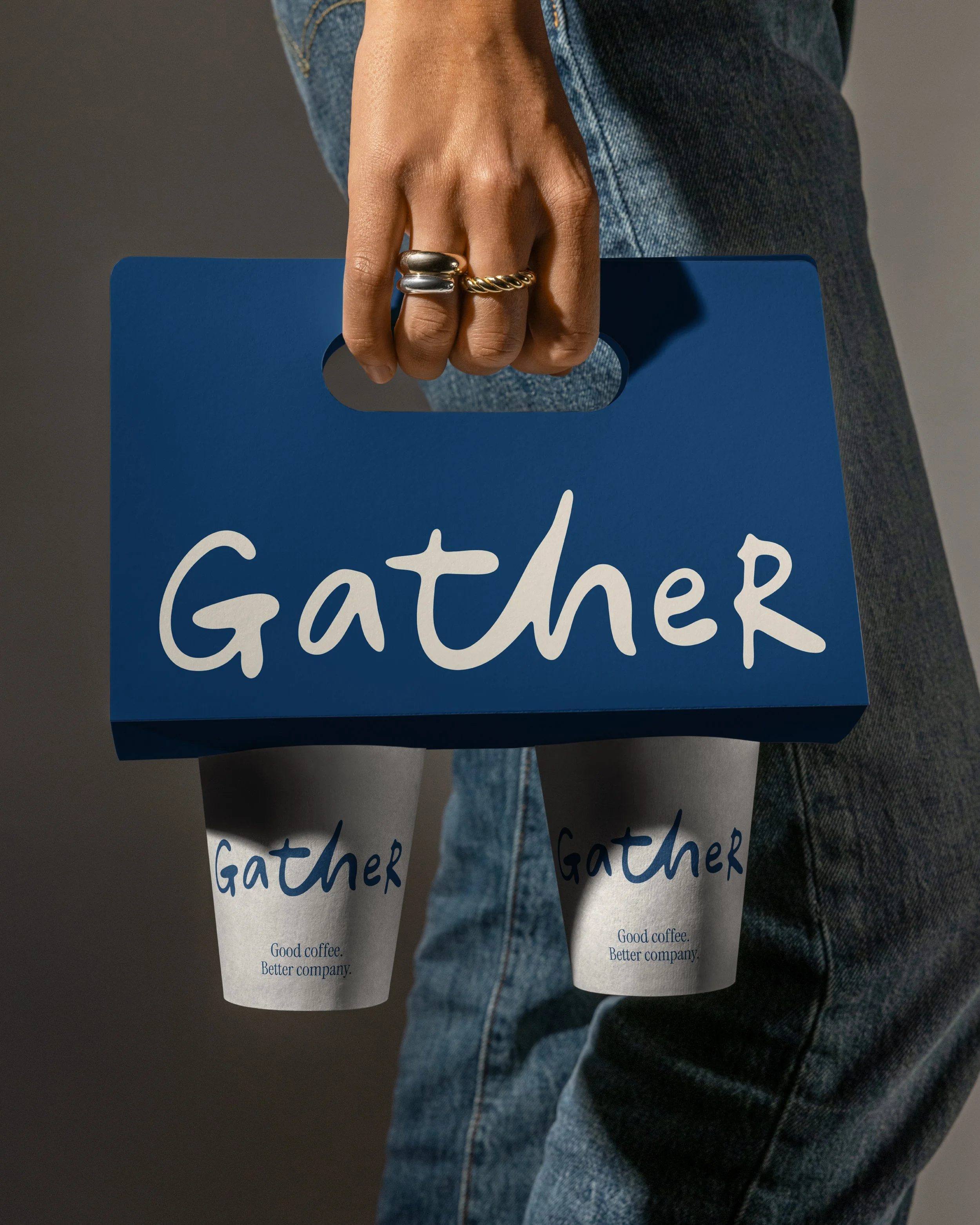A person dressed in blue jeans holding a blue sign that reads 'Gather' in white script. The person has rings on their fingers and is standing against a gray background.