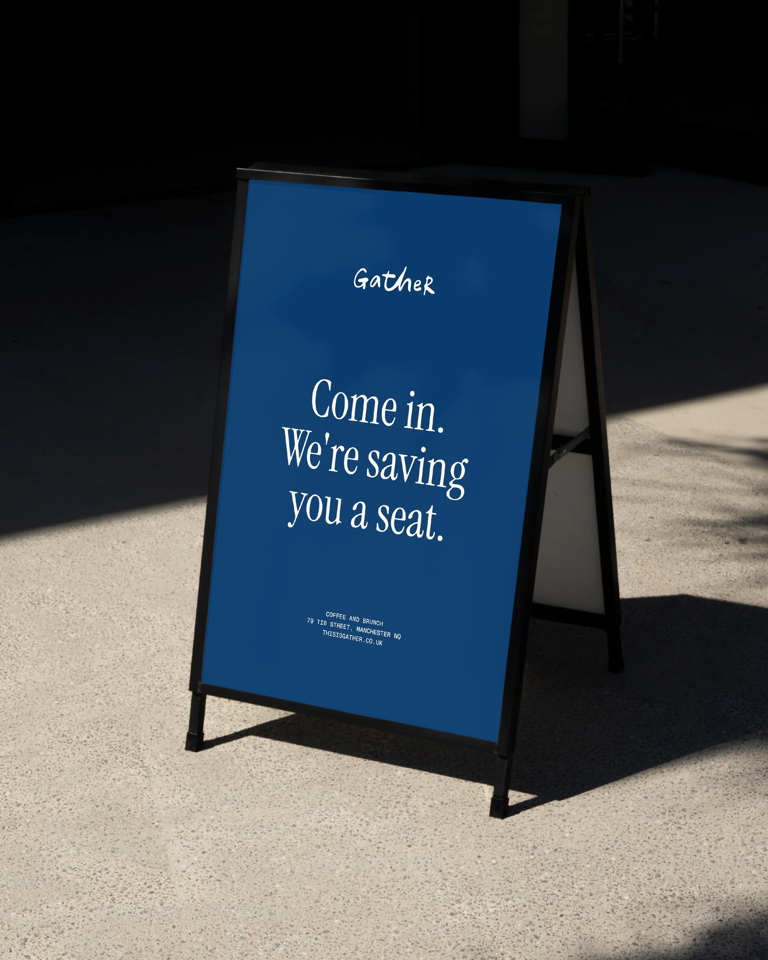 An A-frame signboard on pavement, advertising a coffee and brunch place with the message 'Gather, Come in. We're saying you a seat,' and the address '79 TIB Street, Manchester MQ, thisisgather.co.uk'.