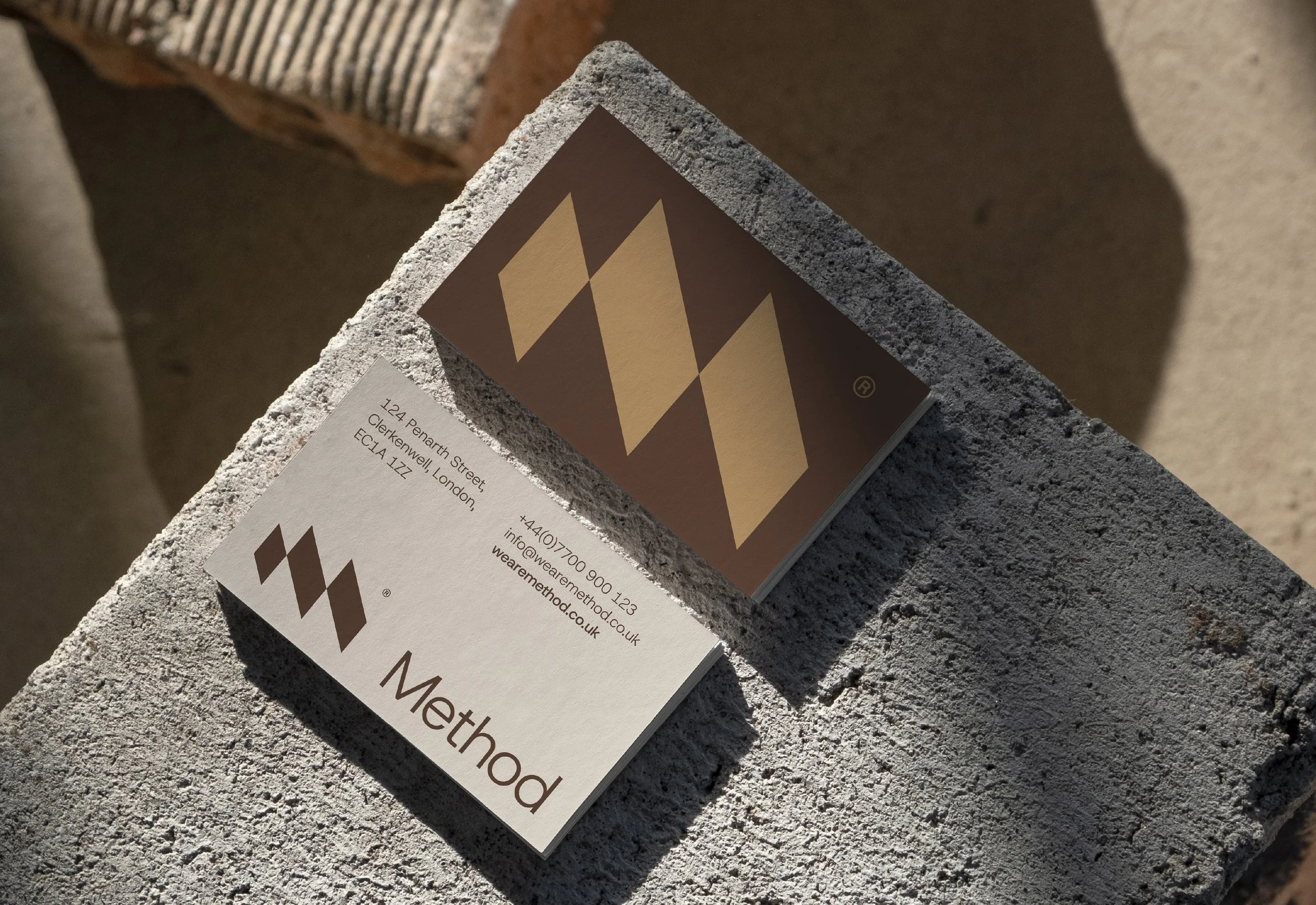Close-up of a business card on a concrete surface with branding, contact information, and a logo featuring two beige diamonds on a brown background.