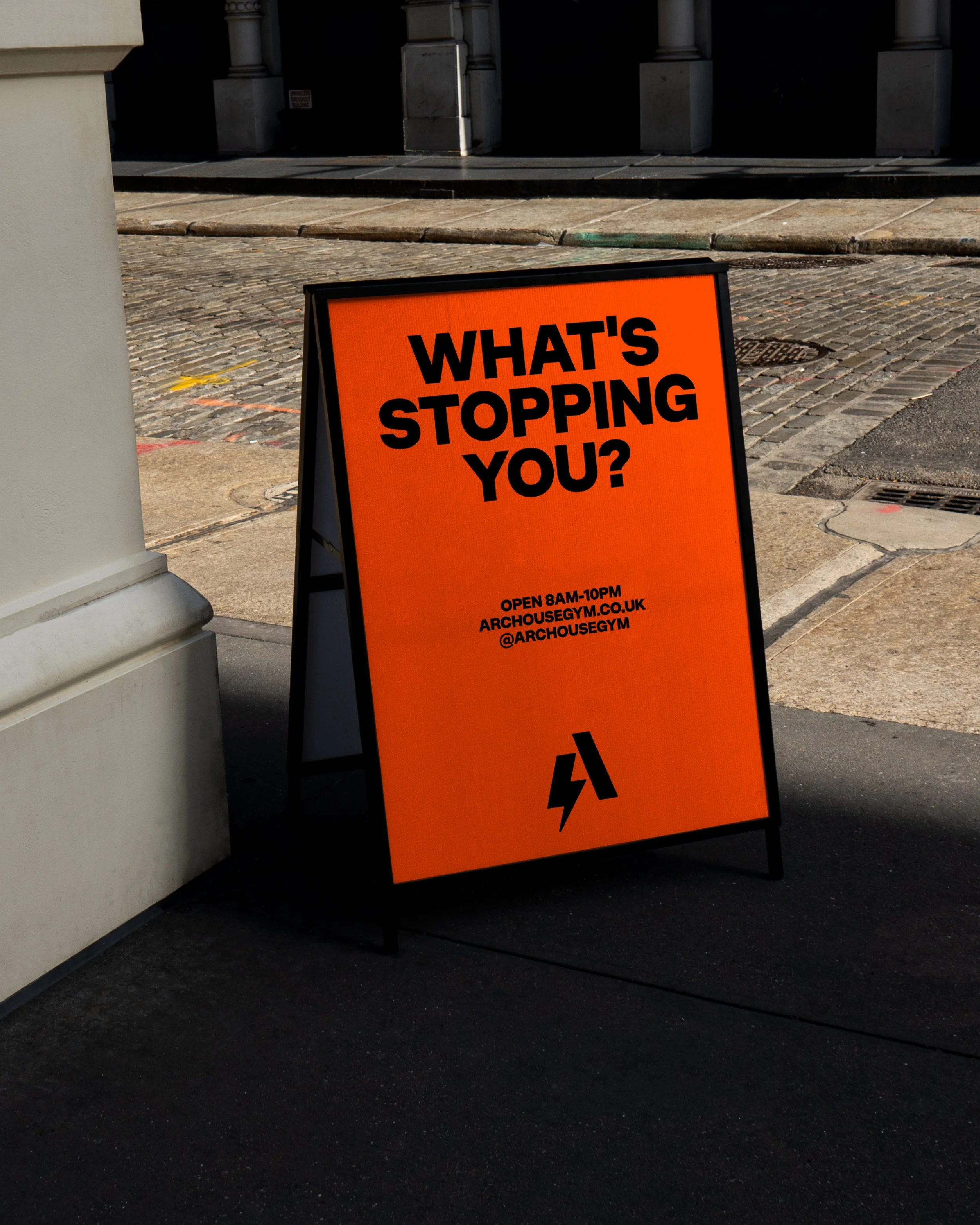 An orange A-frame sidewalk sign with black text that reads 'WHAT'S STOPPING YOU?' alongside additional information about open hours and social media, placed on a city sidewalk near a white pillar and building entrance.