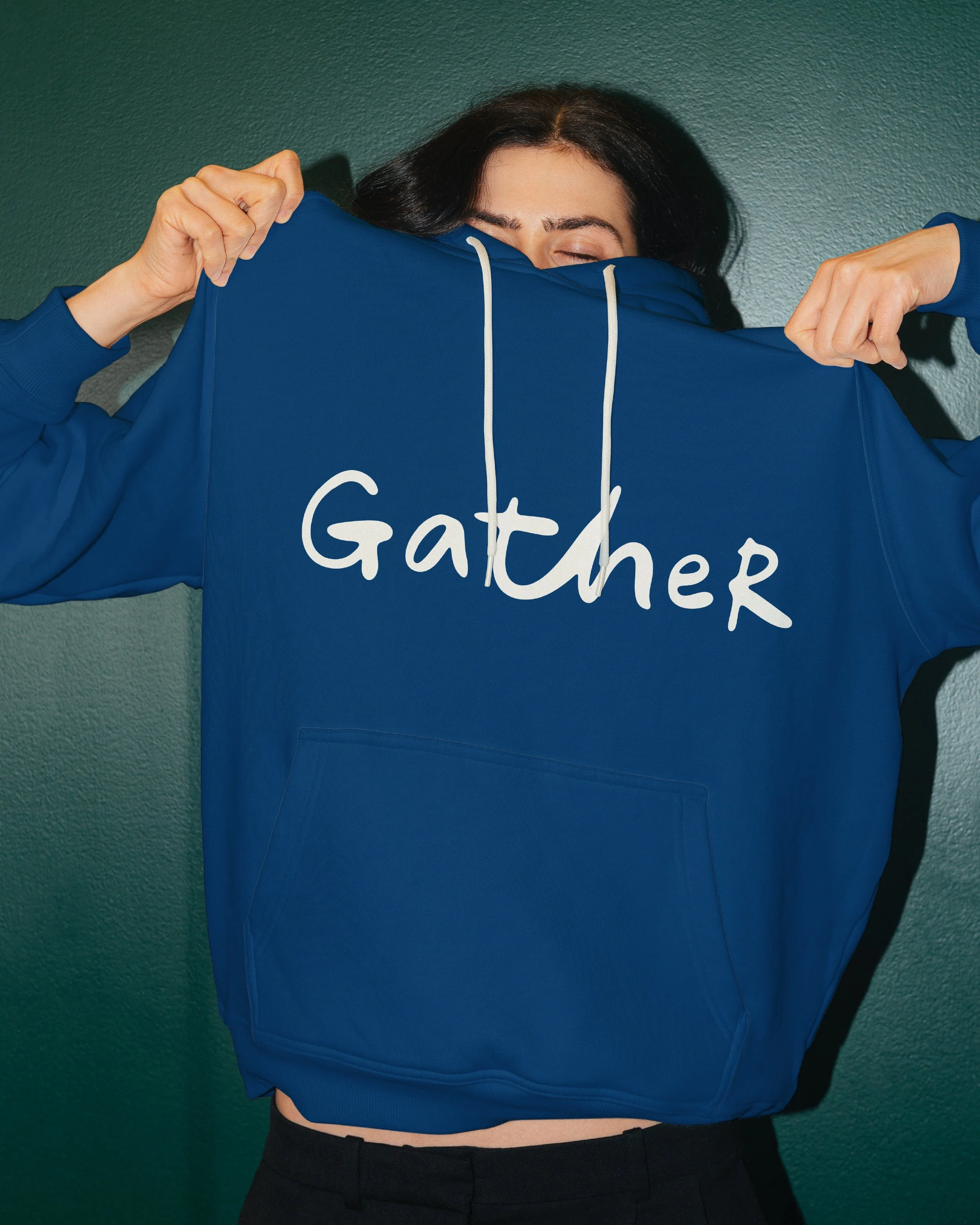 A woman with dark hair is lifting her blue hoodie, revealing the word 'GATHER' written on the front of the hoodie. She is standing against a green textured wall.
