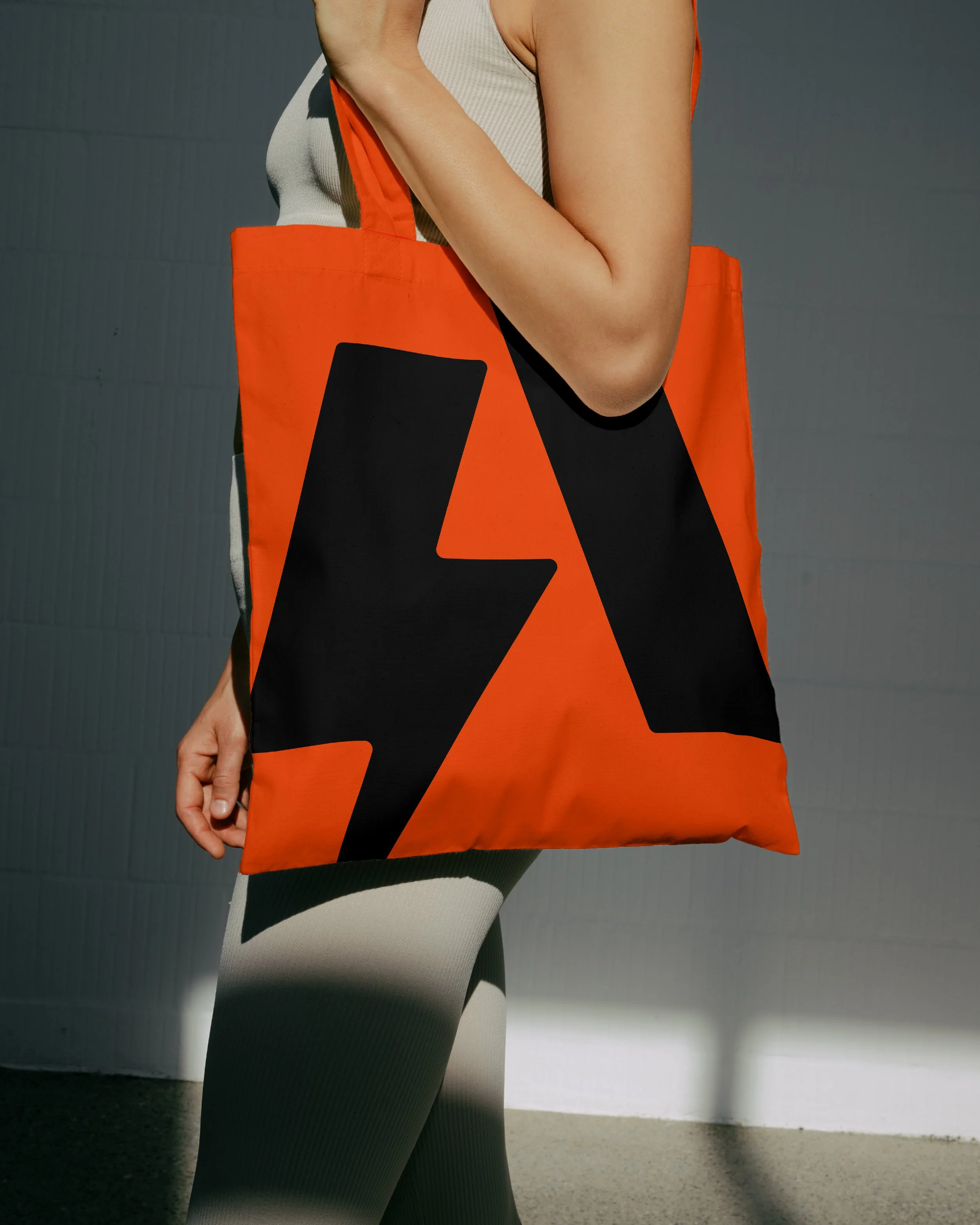 Person holding an orange tote bag with a black abstract design, wearing black and white patterned leggings and a beige sleeveless top, standing in front of a gray wall.