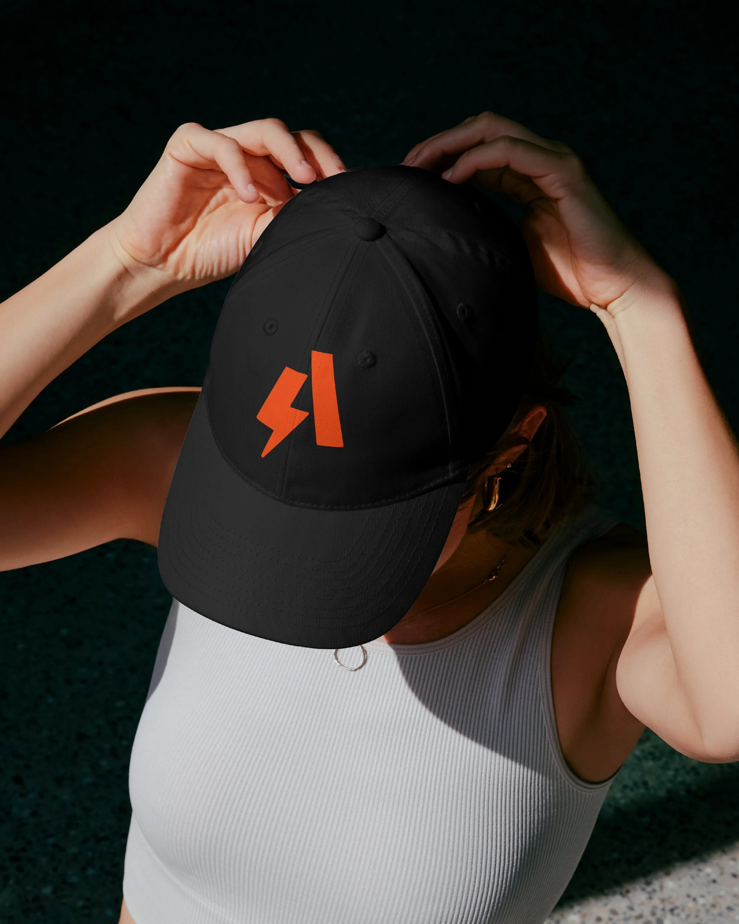 Person adjusting a black cap with an orange logo while sitting on a white surface, wearing a white sleeveless athletic top, with a dark textured background.