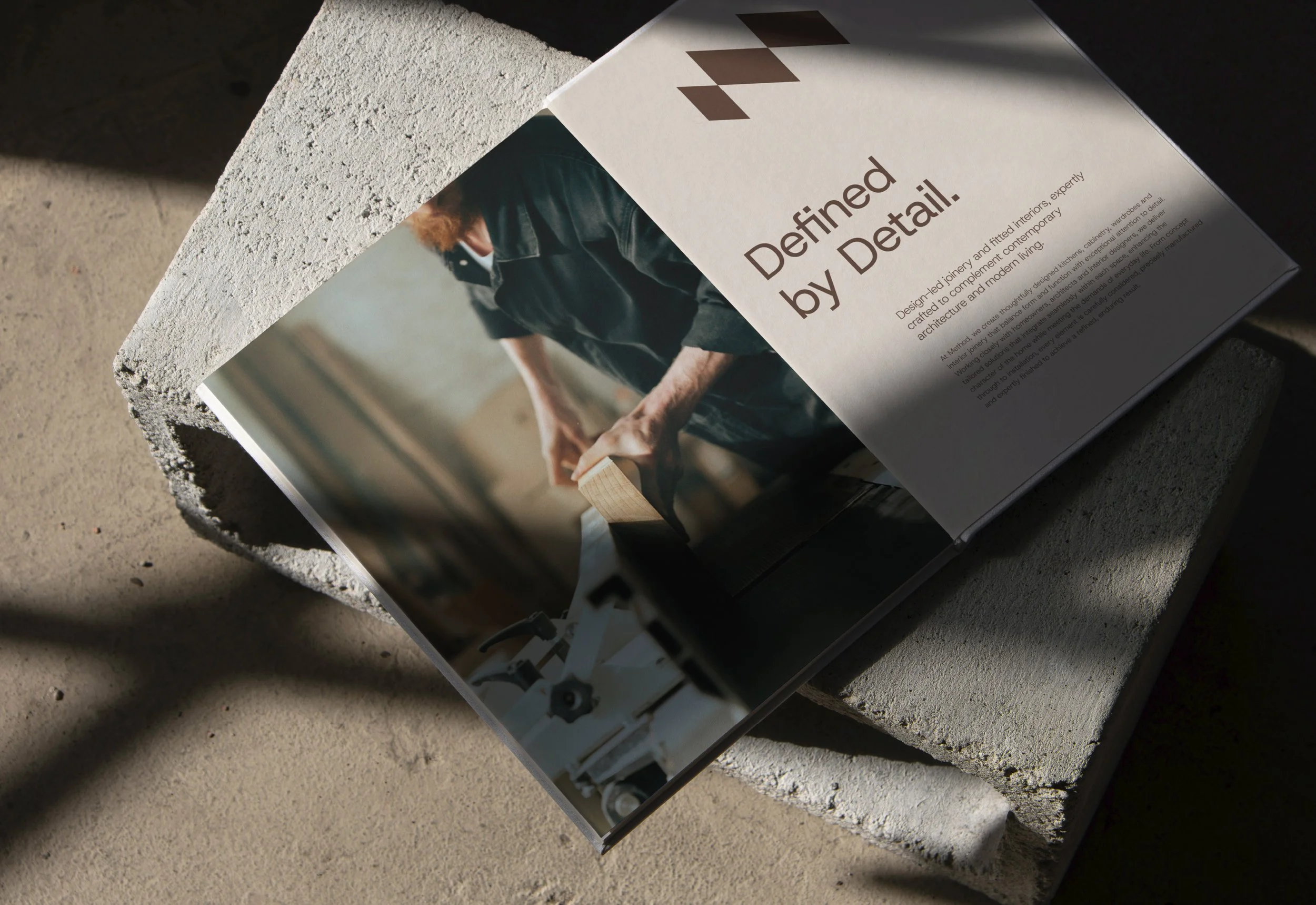 A brochure titled 'Defined by Detail' resting on a rough concrete surface, featuring an image of a person working with wood and design elements related to architecture and craftsmanship.