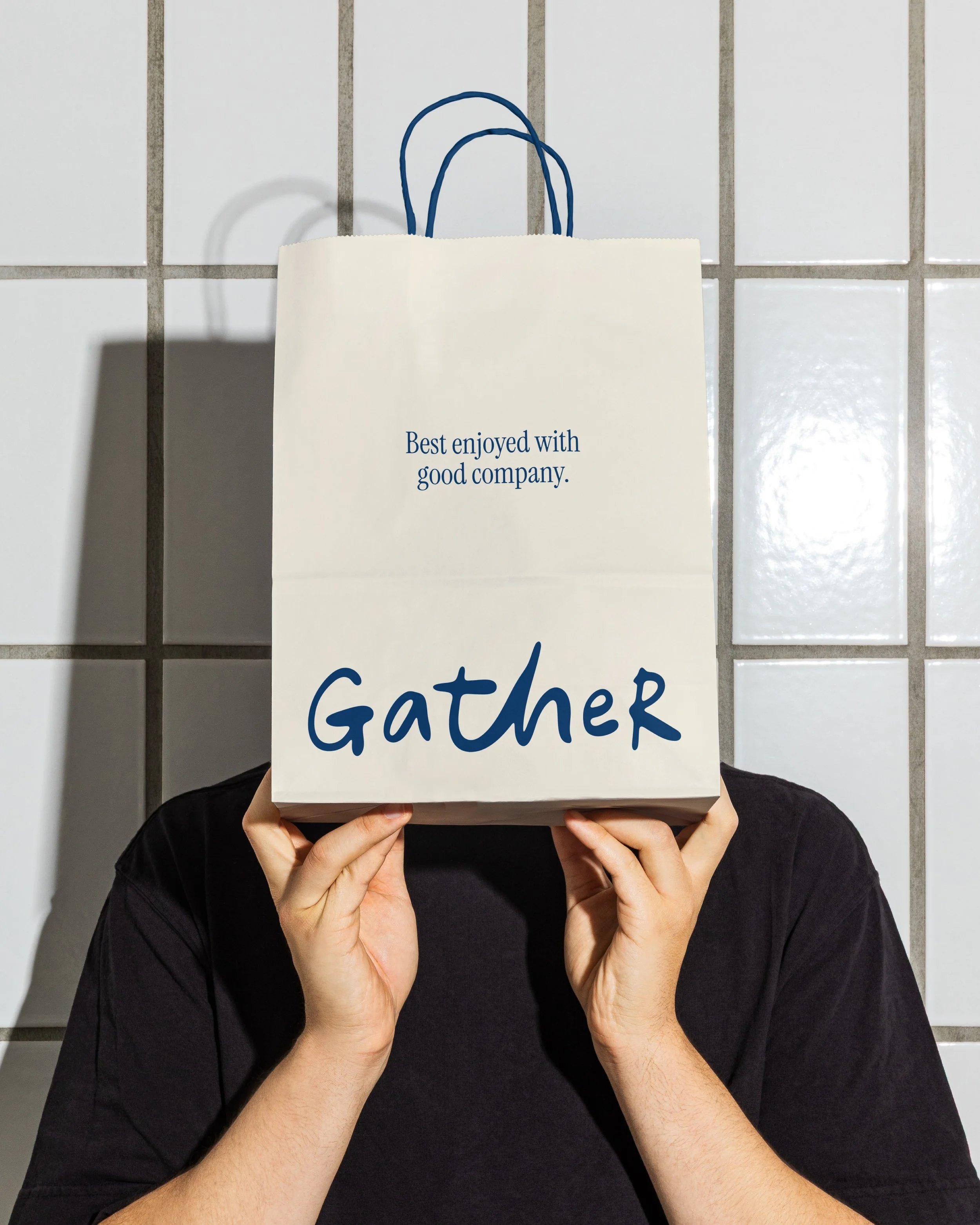 Person holding a white paper bag with blue handles in front of their face, with the text "Best enjoyed with good company" partially visible on the bag, against a tiled wall background.