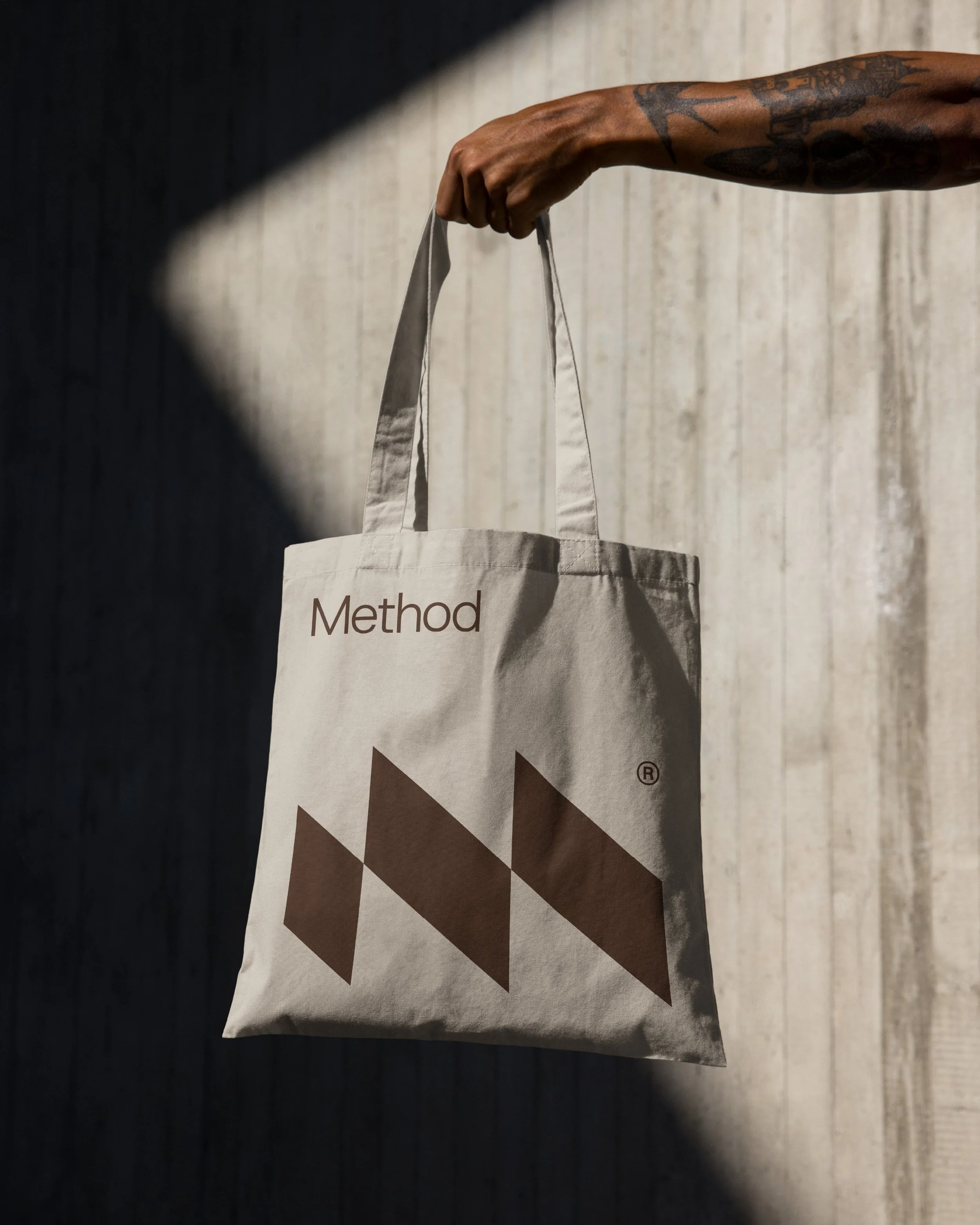A beige tote bag with brown geometric patterns and the word 'Method' printed on it, hanging from a person's hand with tattoos. The background is a light wood wall.