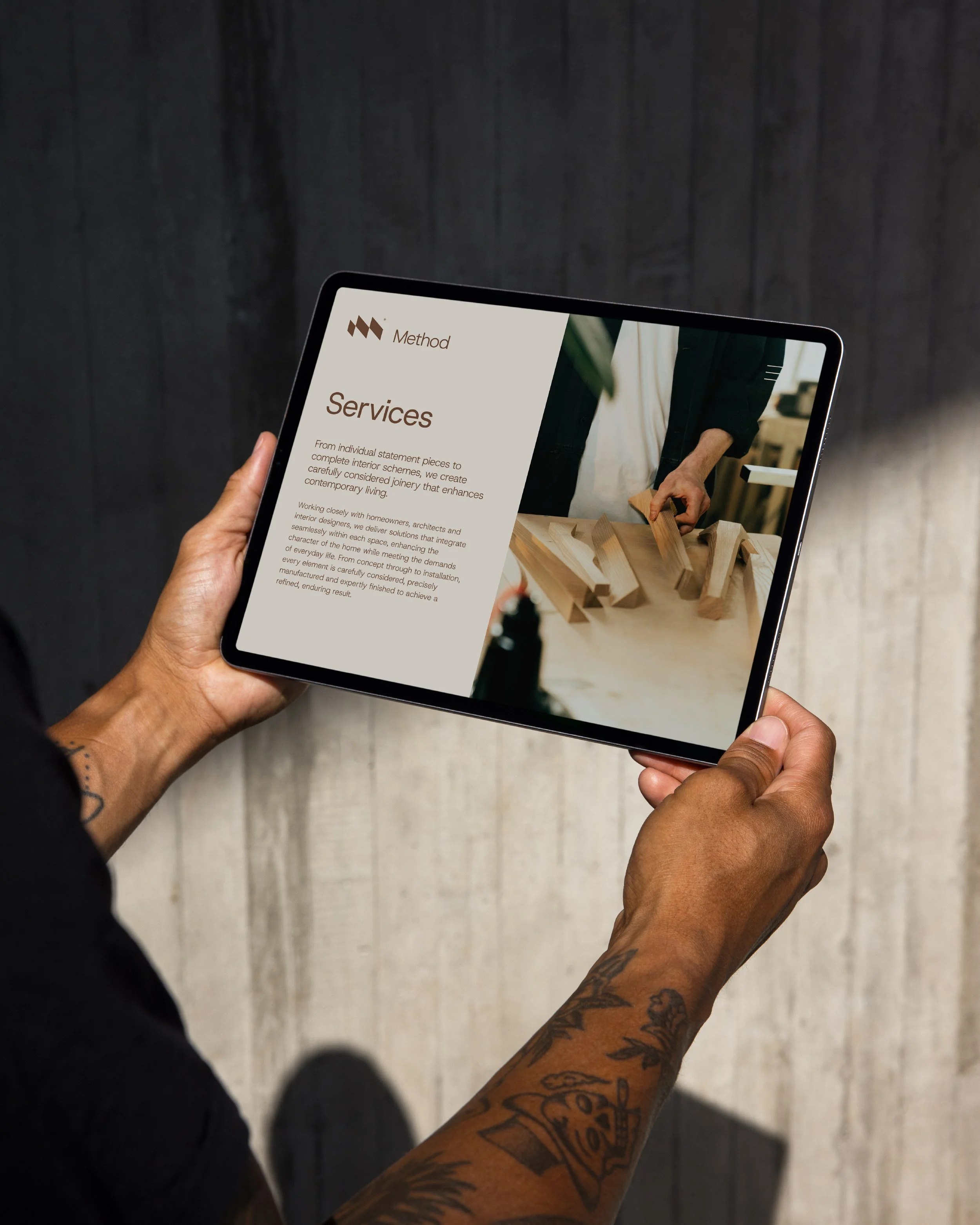 A person holding a tablet displaying a website or document about services. The person has tattoos on their arms and is dressed in dark clothing, standing against a dark wooden background.