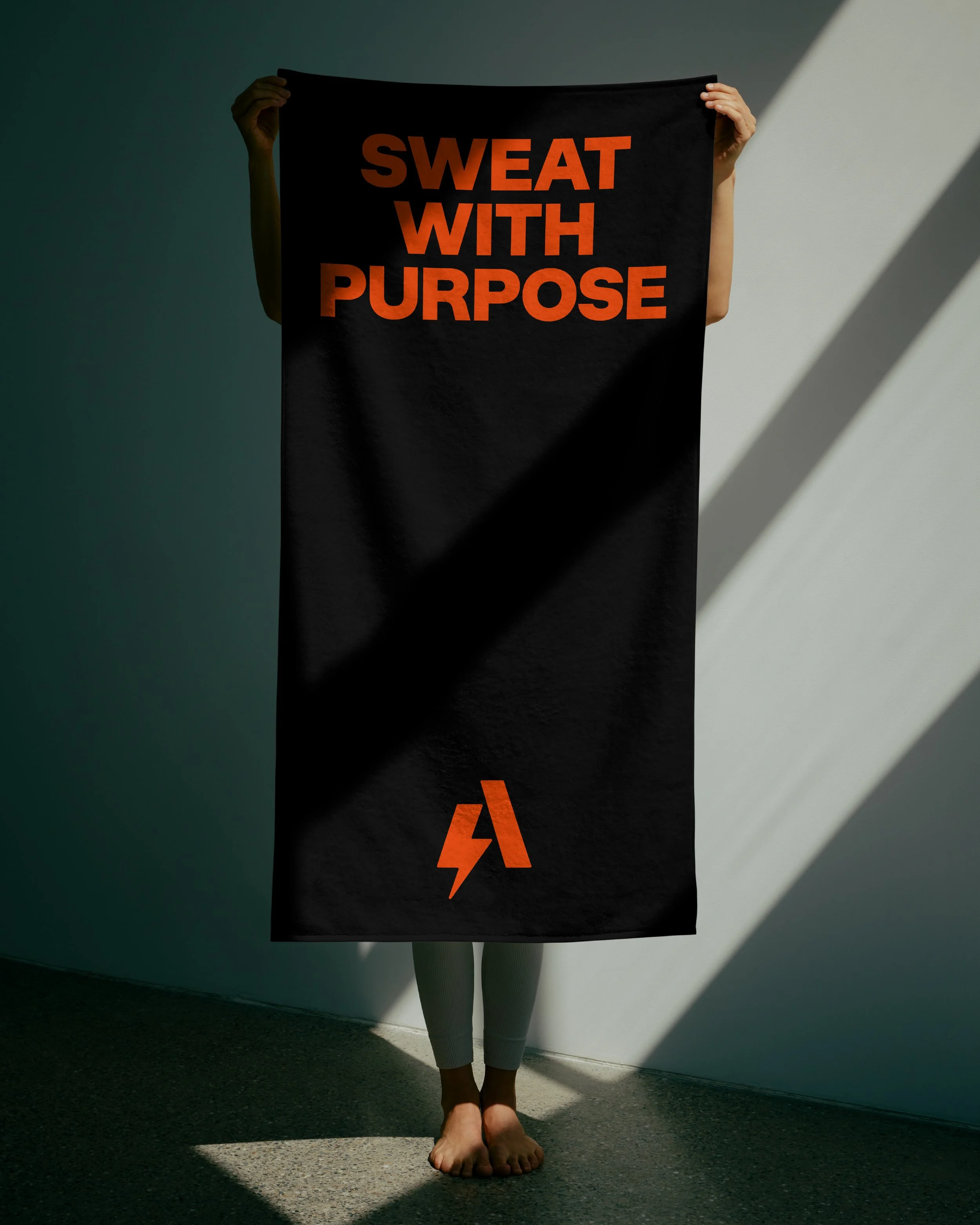 A person holding a black sign with orange text that reads 'SWEAT WITH PURPOSE.' The person is standing barefoot against a white wall with a shadow cast on the wall and floor. The person's face is not visible.
