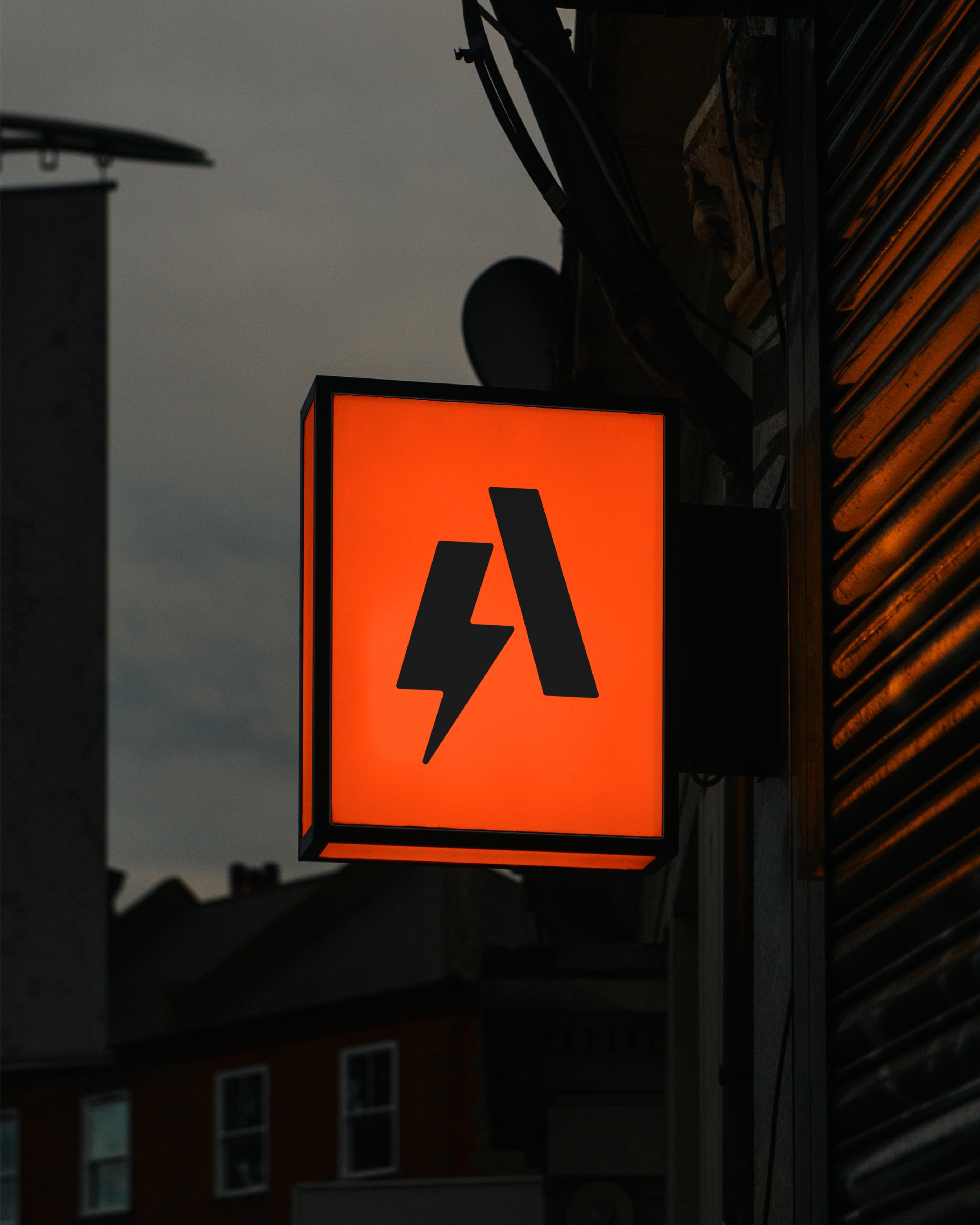 A bright orange warning sign with a black lightning bolt symbol indicating electrical hazard, displayed outside during low light conditions at dusk or evening.