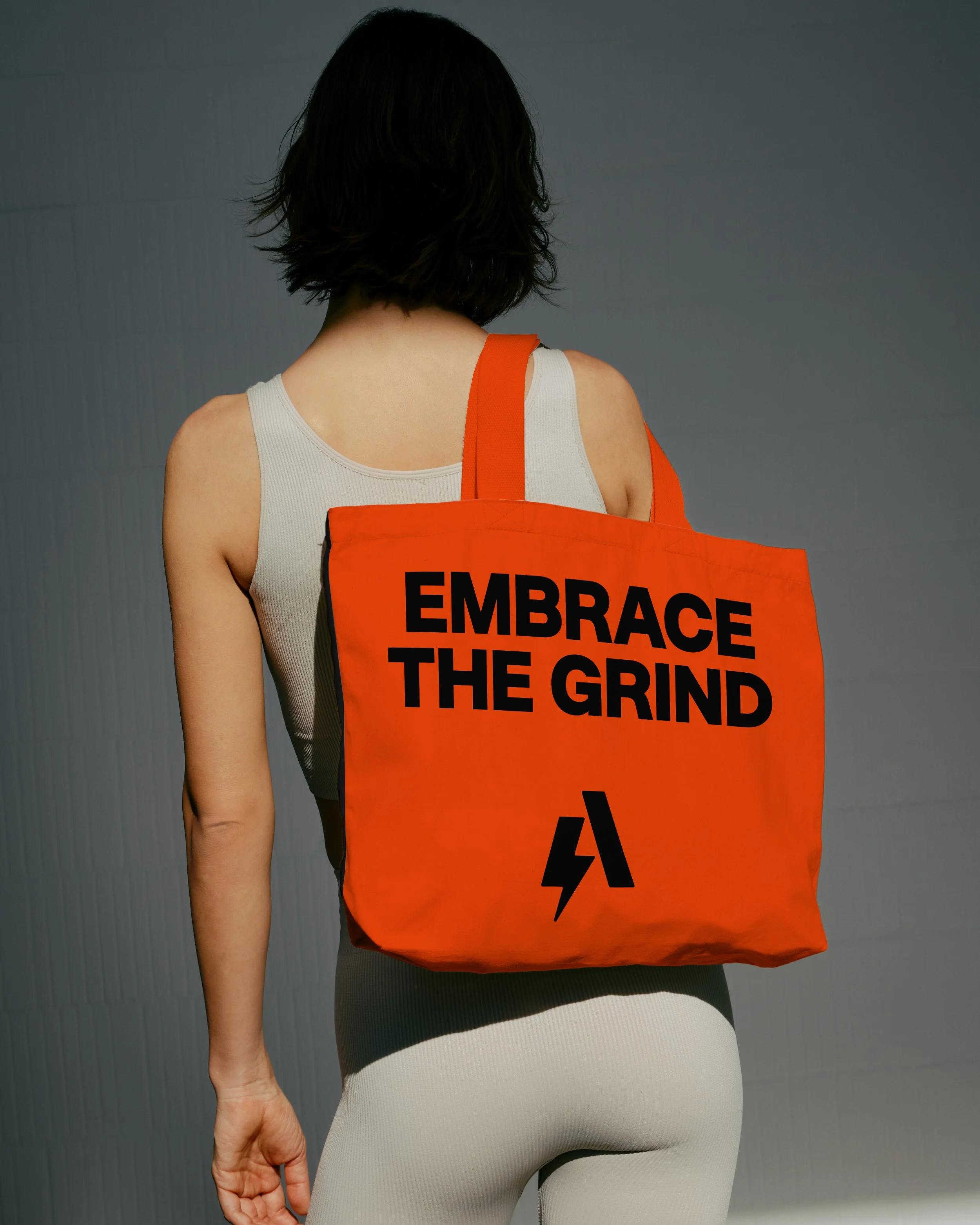 Back view of a woman with dark hair wearing a white tank top and beige pants, carrying an orange bag with black text that reads 'EMBRACE THE GRIND' and a black lightning bolt symbol.