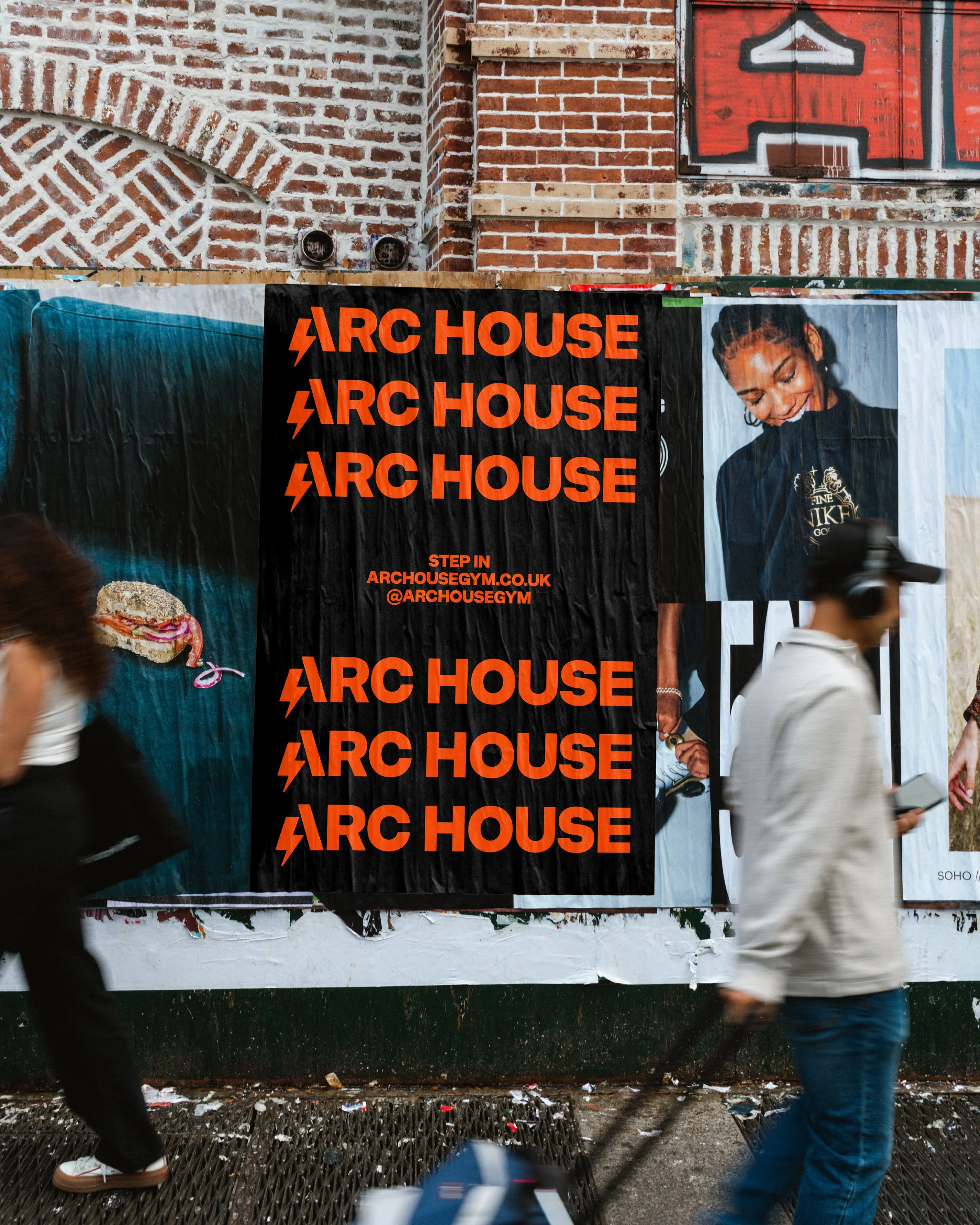 Street scene featuring promotional banners for ARC House gym, a brick building with a graffiti-style red and black arrow symbol, and pedestrians walking past.