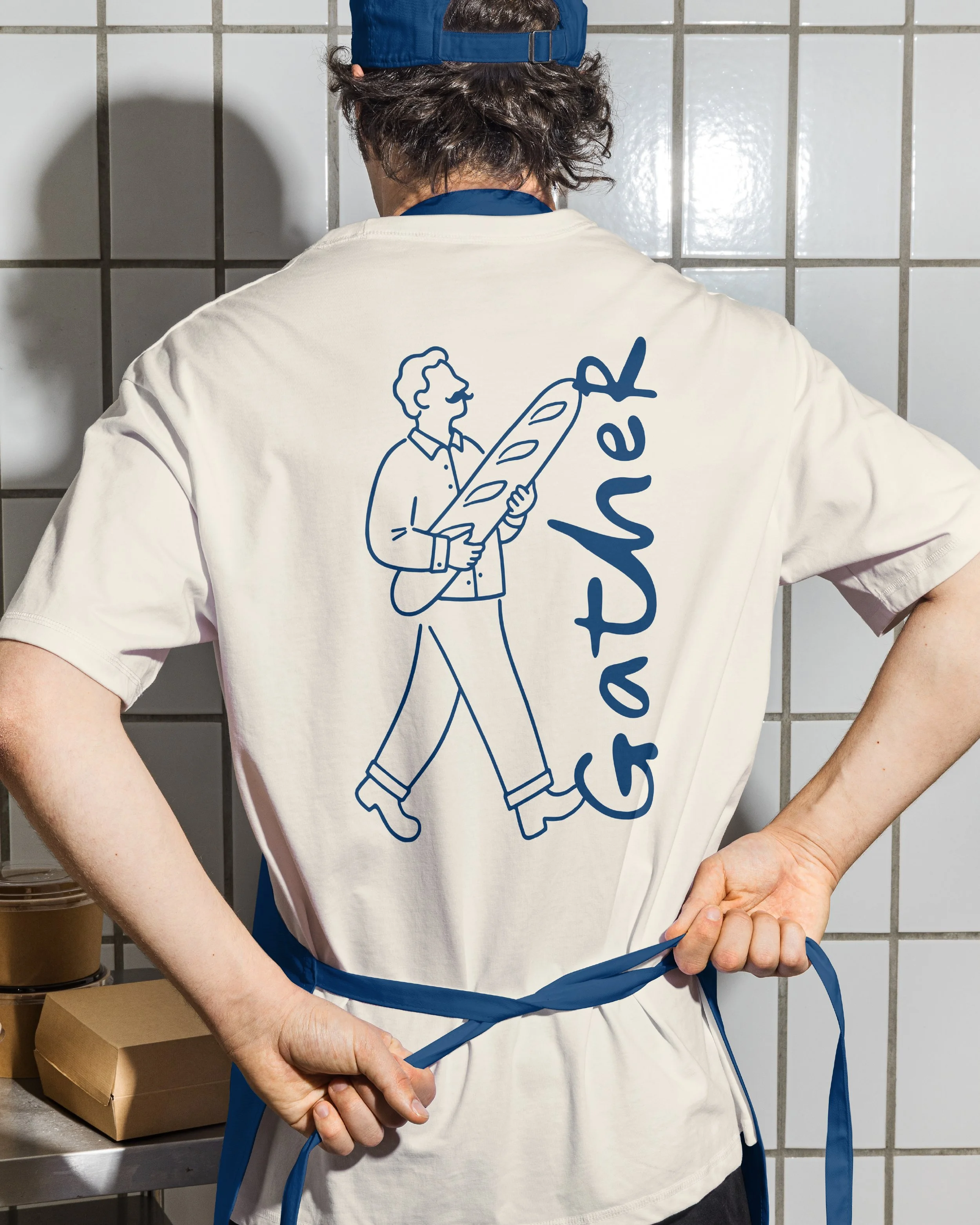 Person wearing a white t-shirt with blue graphic of a man holding a baguette, tying a blue apron in a kitchen or bakery setting.