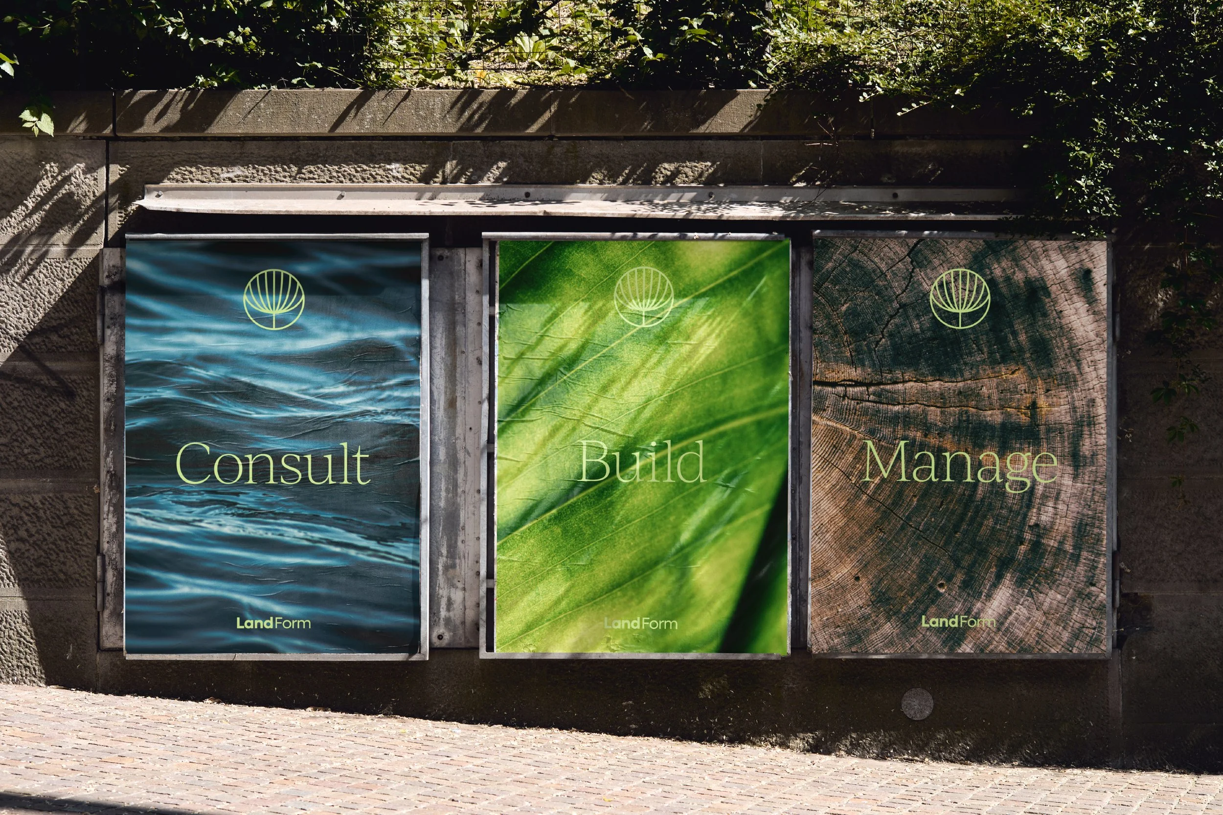 Three outdoor posters on a stone wall with greenery above. The posters have a leaf logo and words 'Consult,' 'Build,' and 'Manage' with the branding 'LandForm' at the bottom of each.