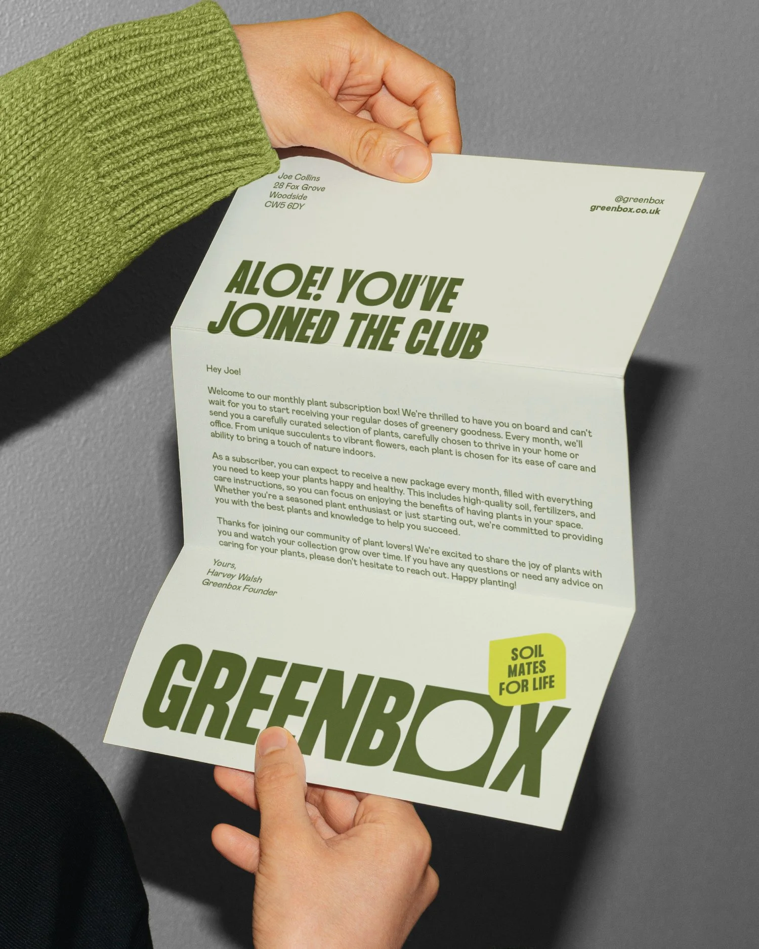 A person holding a Greenbox flyer promoting plant subscription with bold text 'Aloe! You've Joined the Club' and 'Greenbox' logo with the tagline 'Soil Mates for Life'.