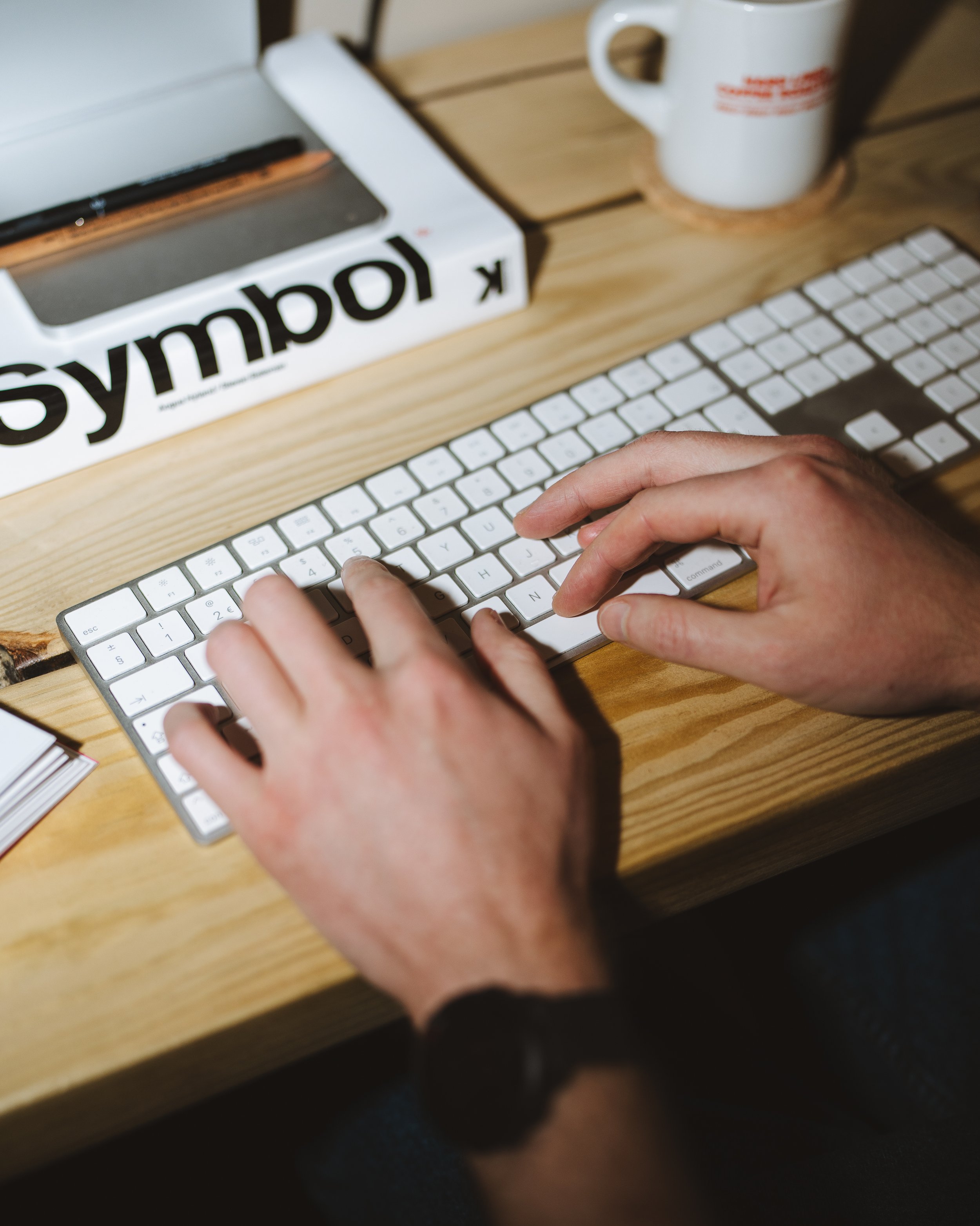 Person typing on a white keyboard at a wooden desk, with a white mug and a white box labeled 'symbol' nearby.