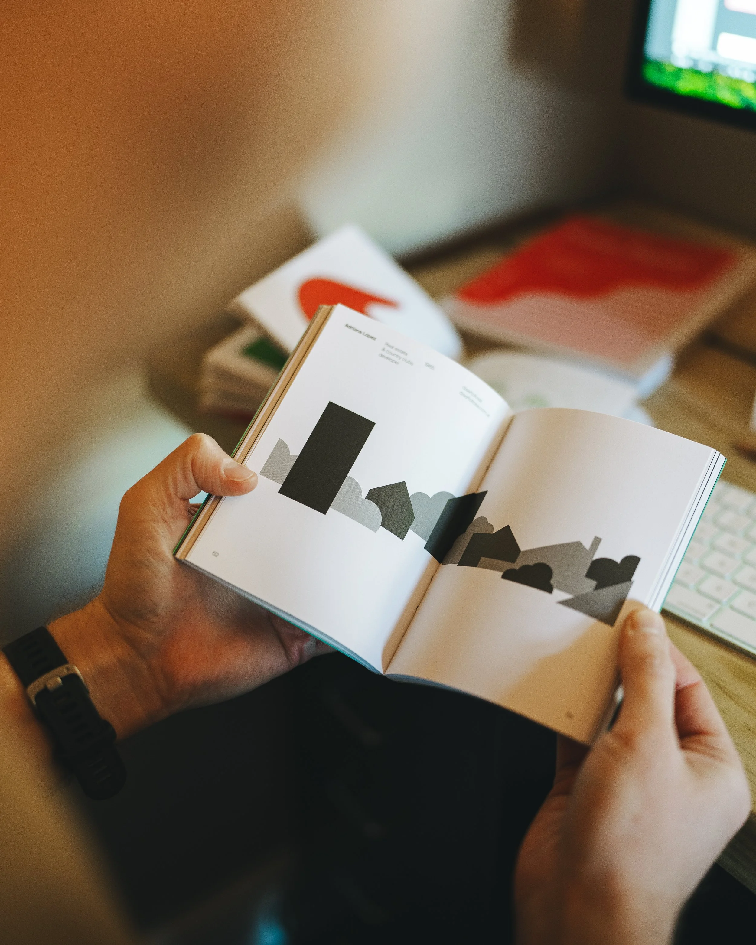 Person holding an open book with black and gray abstract shapes on the pages, with a computer monitor and some more books in the background.
