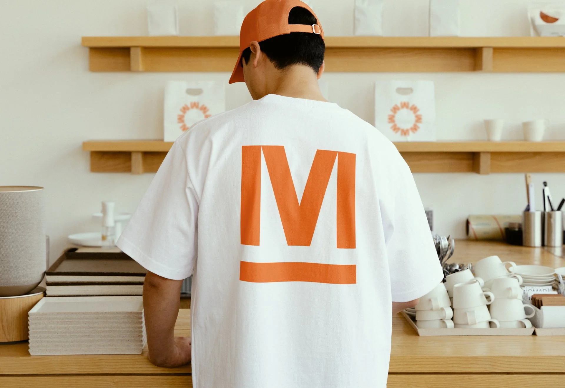 Back of a person wearing a white T-shirt with orange 'M' and underline, standing behind a counter showing various mugs, plates, and shelves with packaging in a minimalist cafe or store.