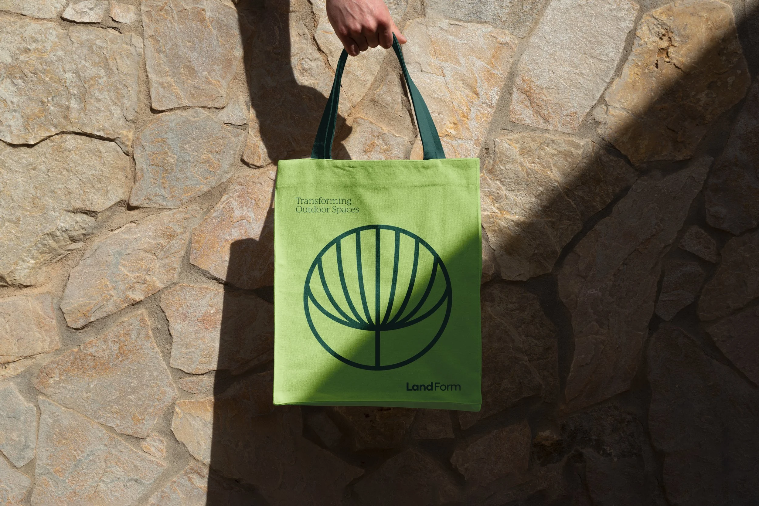 Green tote bag with black line drawing of a stylized leaf or shell, held up by a person against a stone wall background.