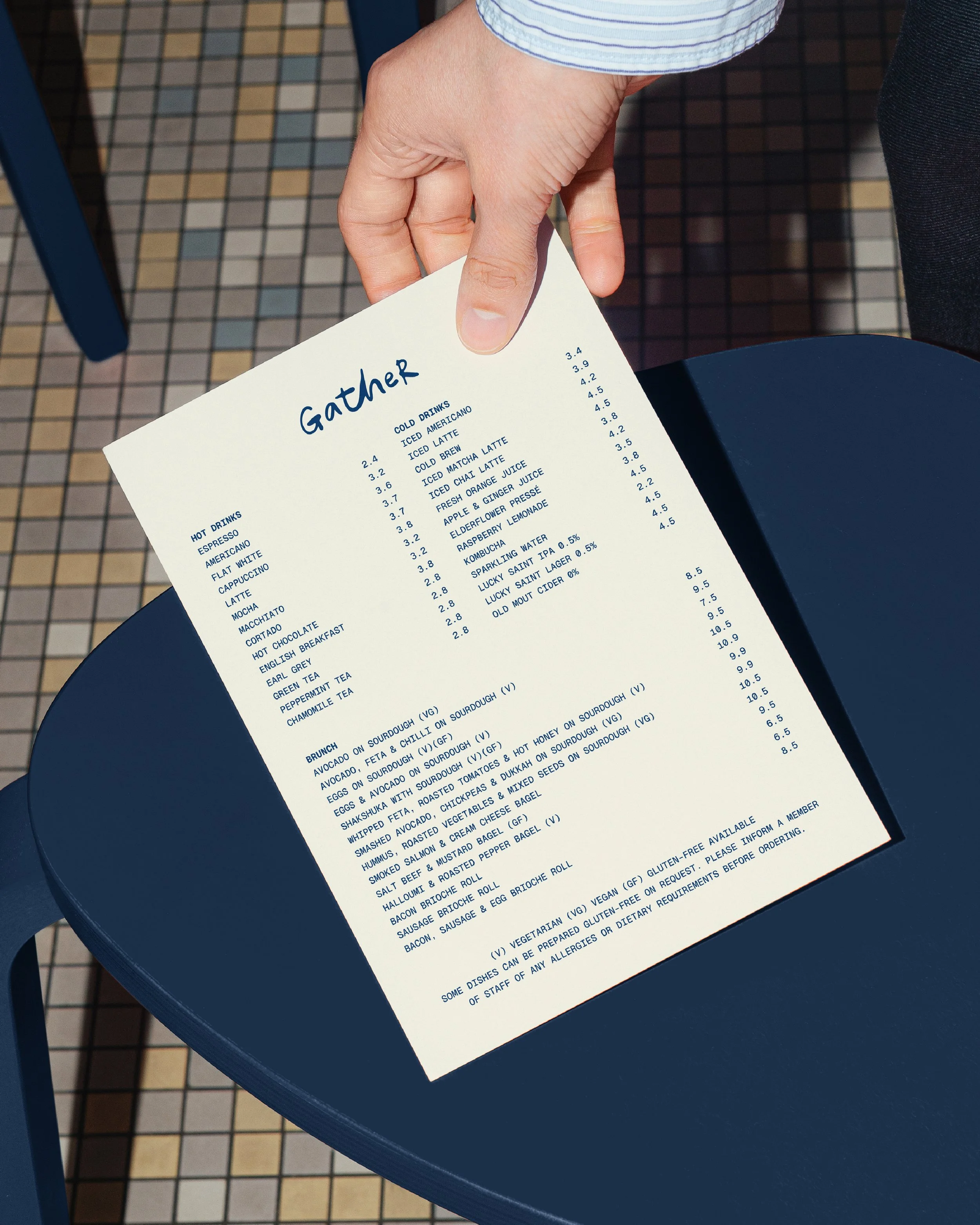 Close-up of a person holding a white menu with blue text at a restaurant or café; the menu lists various drinks and dishes.