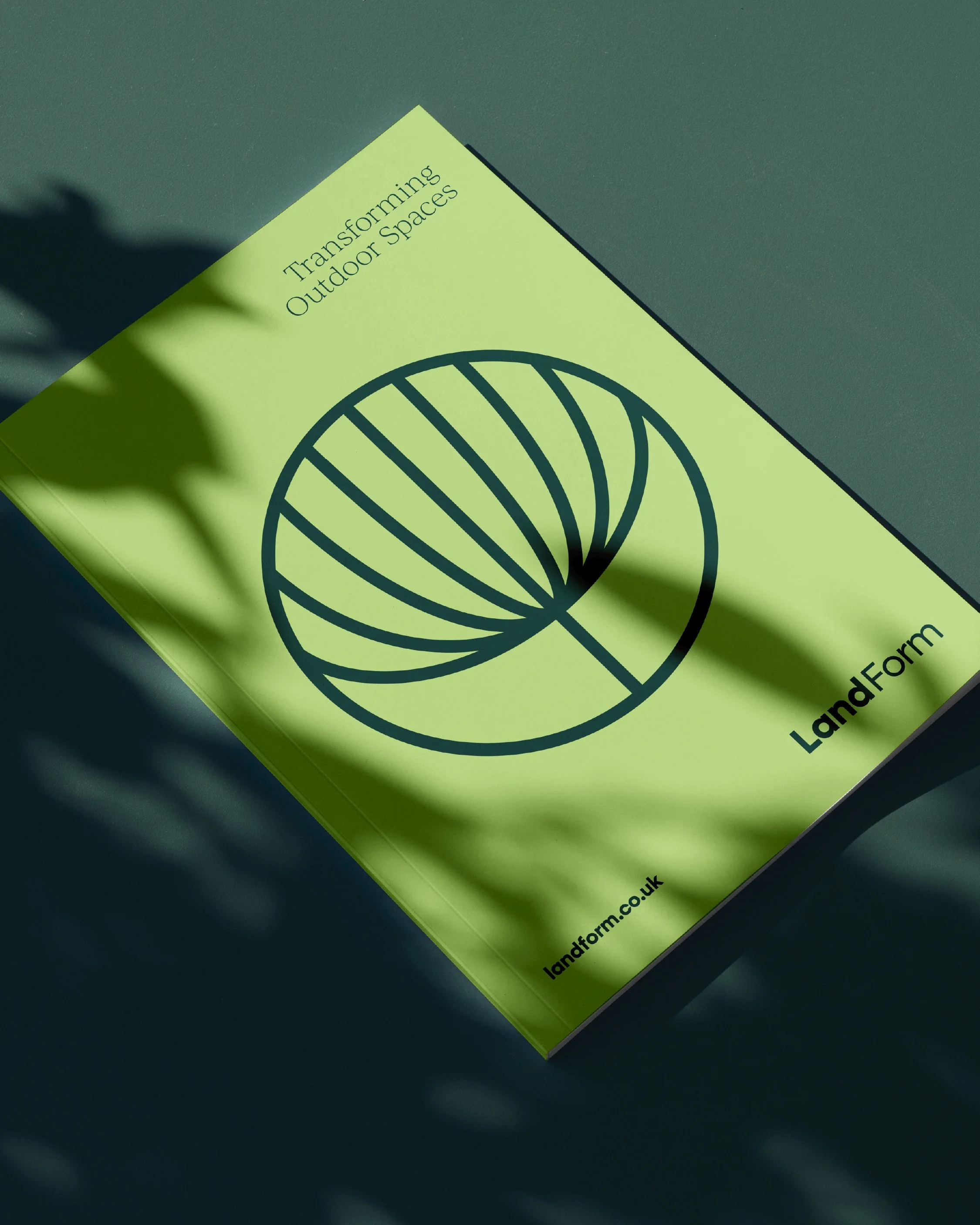 Green brochure titled 'Transforming Outdoor Spaces' with a black abstract spiral design and the logo 'LandForm' at the bottom right, on a dark surface with shadowed green leaves.