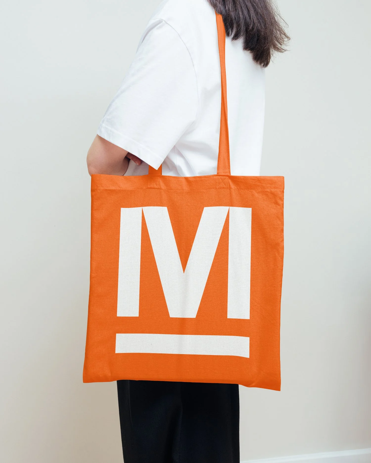 Person holding an orange tote bag with a large white 'M' on it, wearing a white shirt.