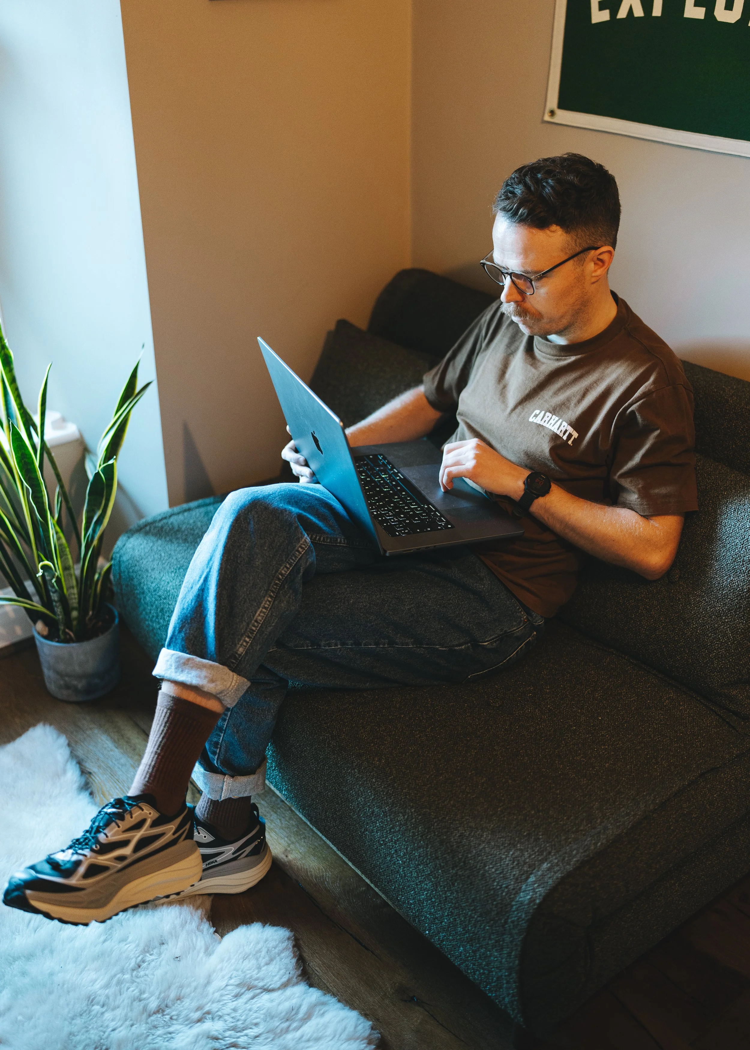 A man with glasses, wearing a brown t-shirt, jeans, and sneakers, sitting on a dark gray sofa, working on a laptop. There's a plant in a pot and a white furry rug nearby, with a wall featuring a green sign in the background.