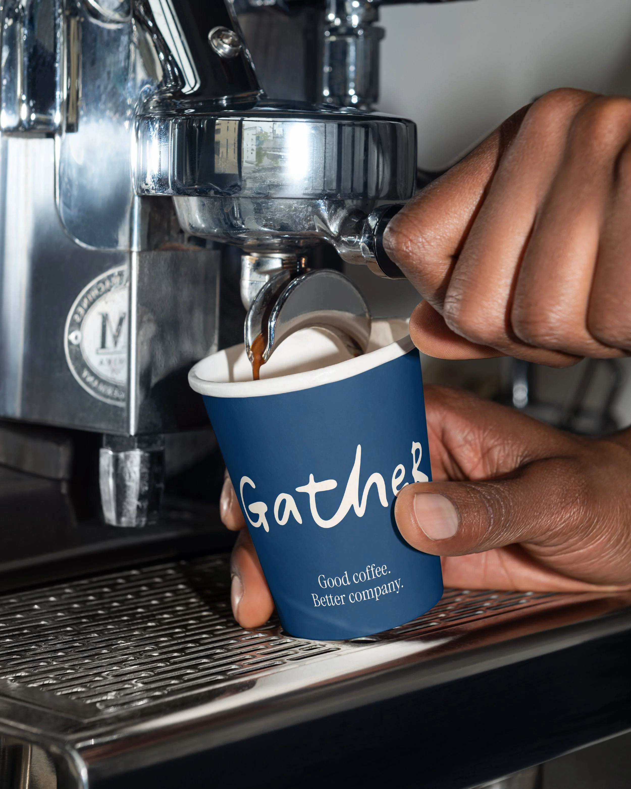 A barista is using a coffee machine to pour brewed coffee into a blue disposable cup with white text that reads 'GATHER.' The coffee machine is metallic with visible knobs and a spout. The person's hand is holding the cup under the spout to collect the coffee.