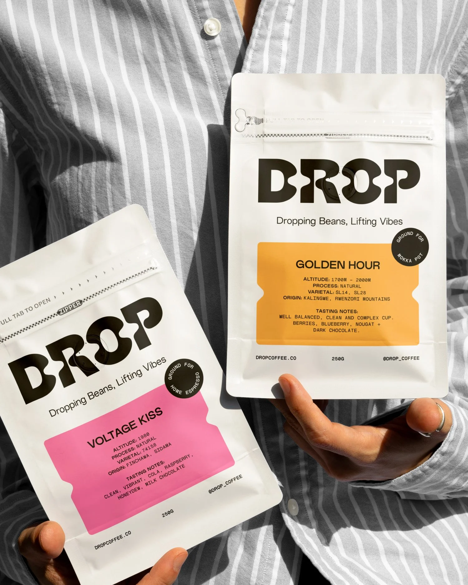 Two bags of Drop coffee with different flavors, one labeled 'Voltage Kiss' with pink label and the other labeled 'Golden Hour' with orange label, held by a person wearing a gray striped shirt.