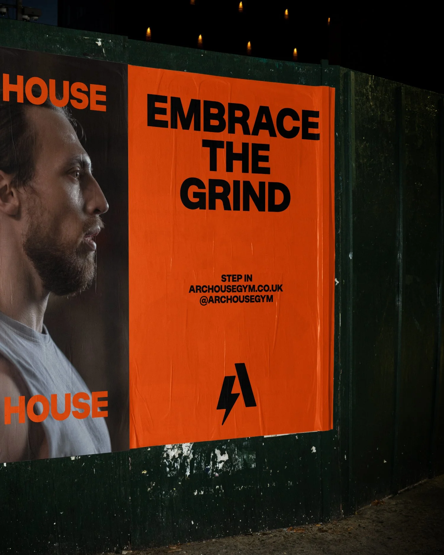 A large orange banner with black text and a photo of a man on the left side. The text reads "EMBRACE THE GRIND" and includes website and social media information for archousegym.co.uk and @archousegym. The banner is mounted on a dark green fence.