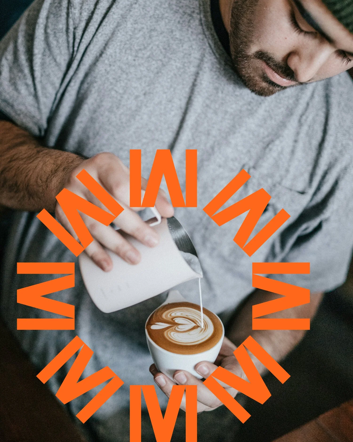 A man in a gray t-shirt is pouring steamed milk into a cup of coffee, creating latte art on top.