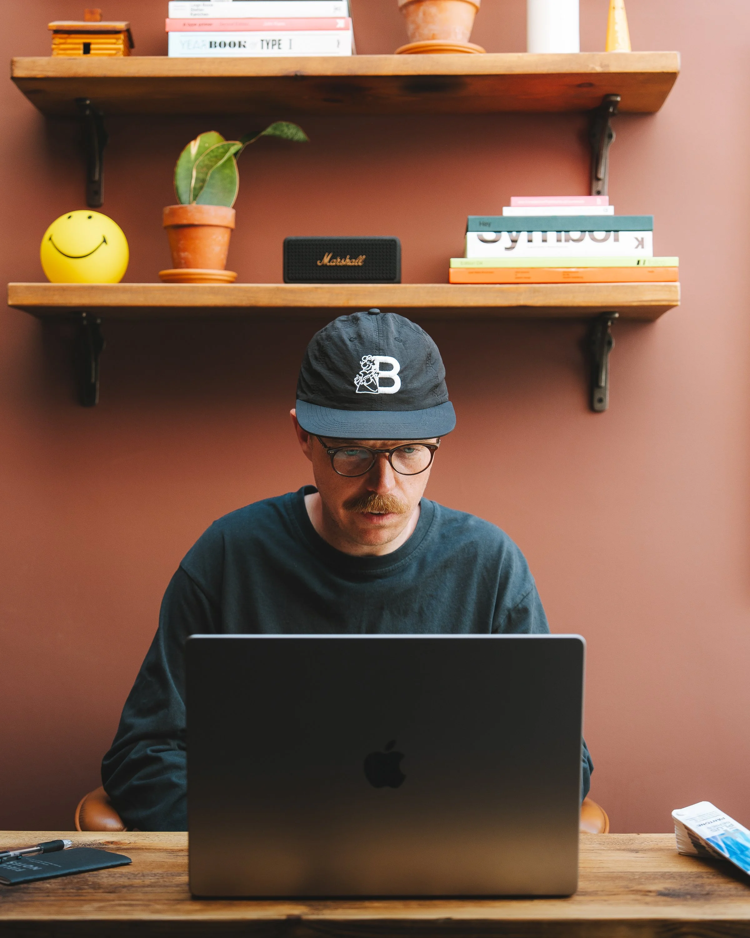 A man wearing glasses and a black cap with a white 'B' on it, sitting at a wooden desk and working on a MacBook laptop. Behind him are two wooden shelves mounted on a reddish-brown wall, holding several books, a small potted plant, a Marshall speaker, and a yellow smiley face ball. There are also various objects on the desk, including a pen and a box of tissues.