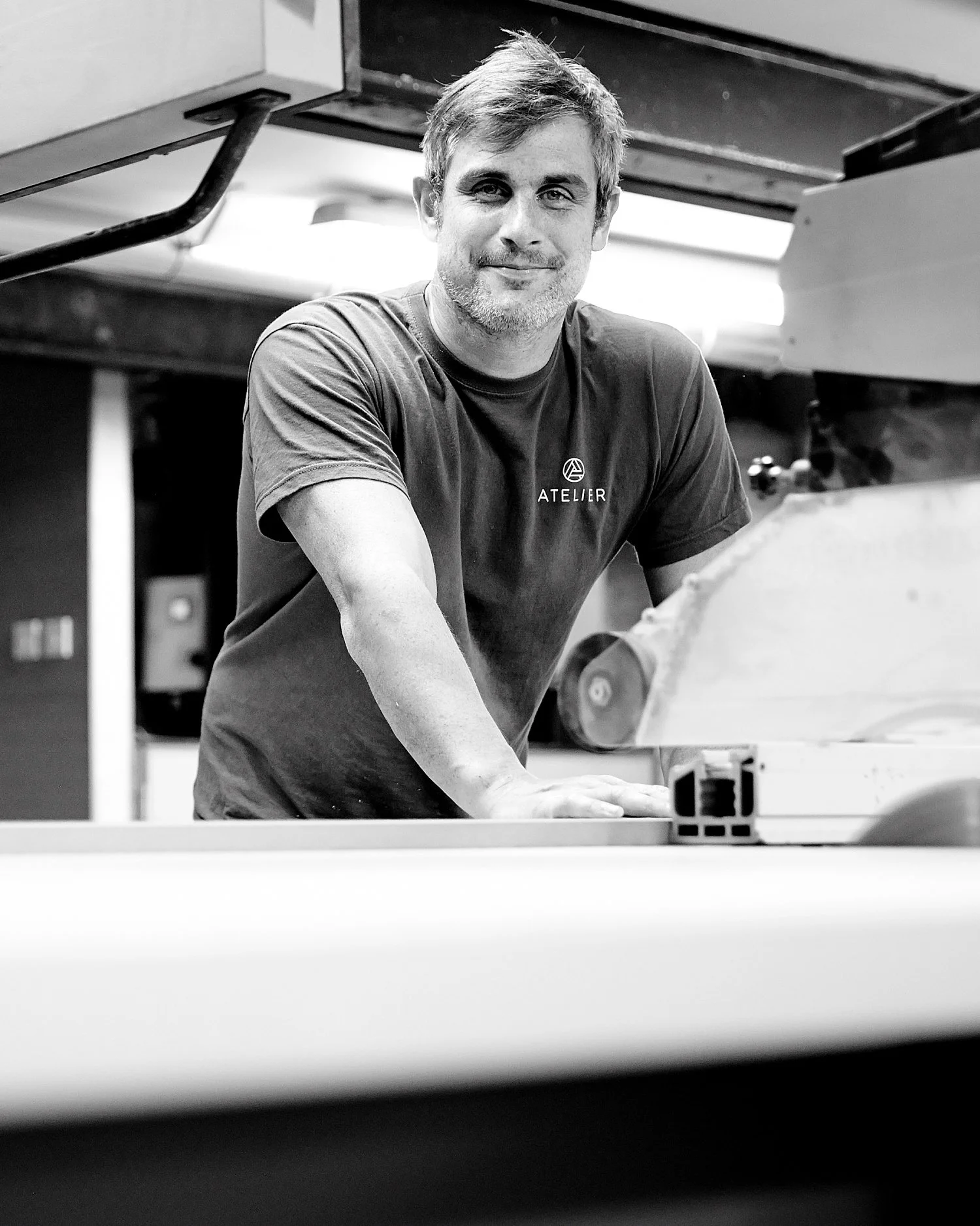 A man with short hair and a beard, wearing a T-shirt with the word 'ATELIER' on it, leaning on a workbench in a workshop, smiling at the camera. There is a saw in the foreground.