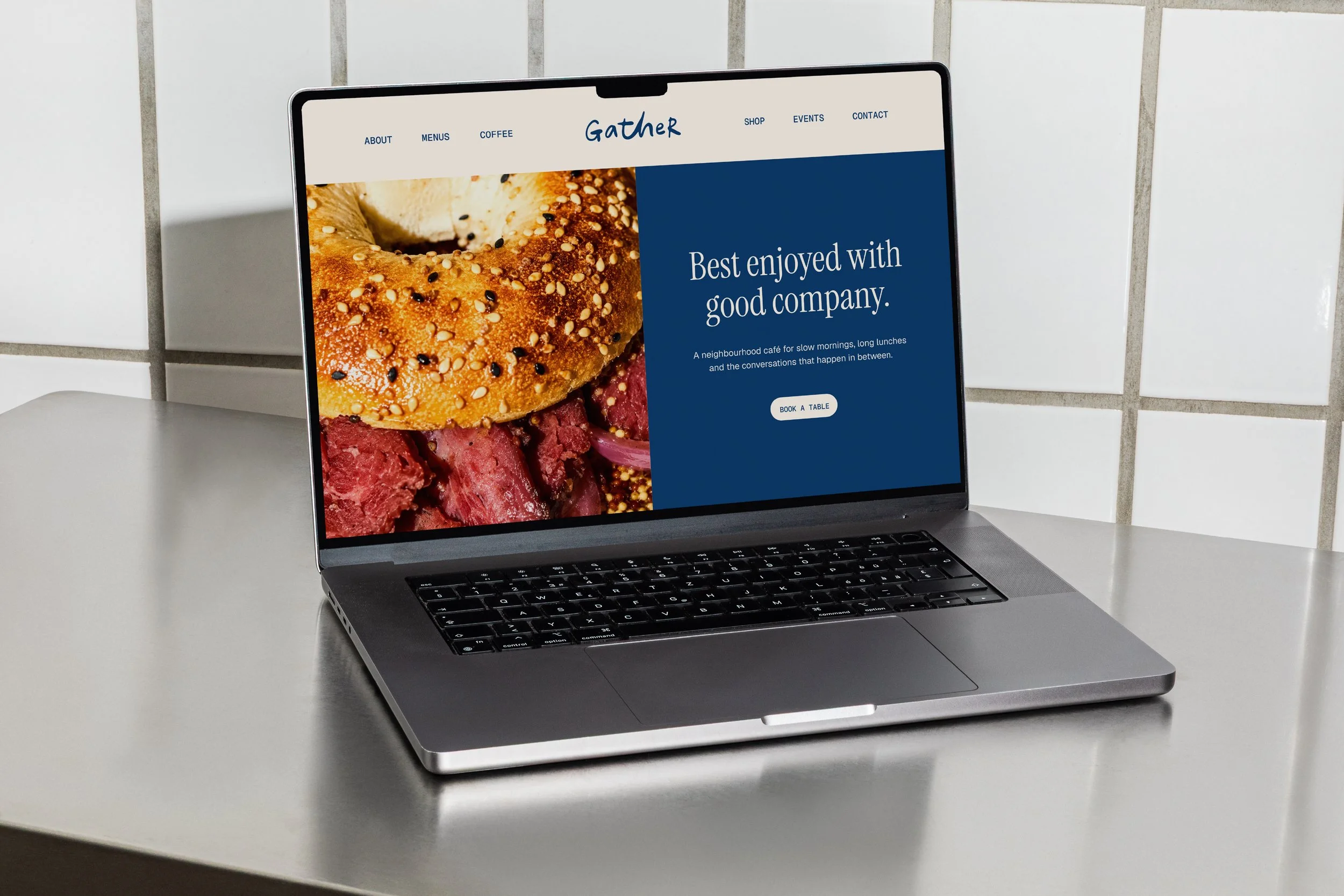 Laptop displaying a website for a café named 'Gather' with an image of a bagel with sesame seeds, alongside text about enjoying good company, on a white table against a tiled wall background.