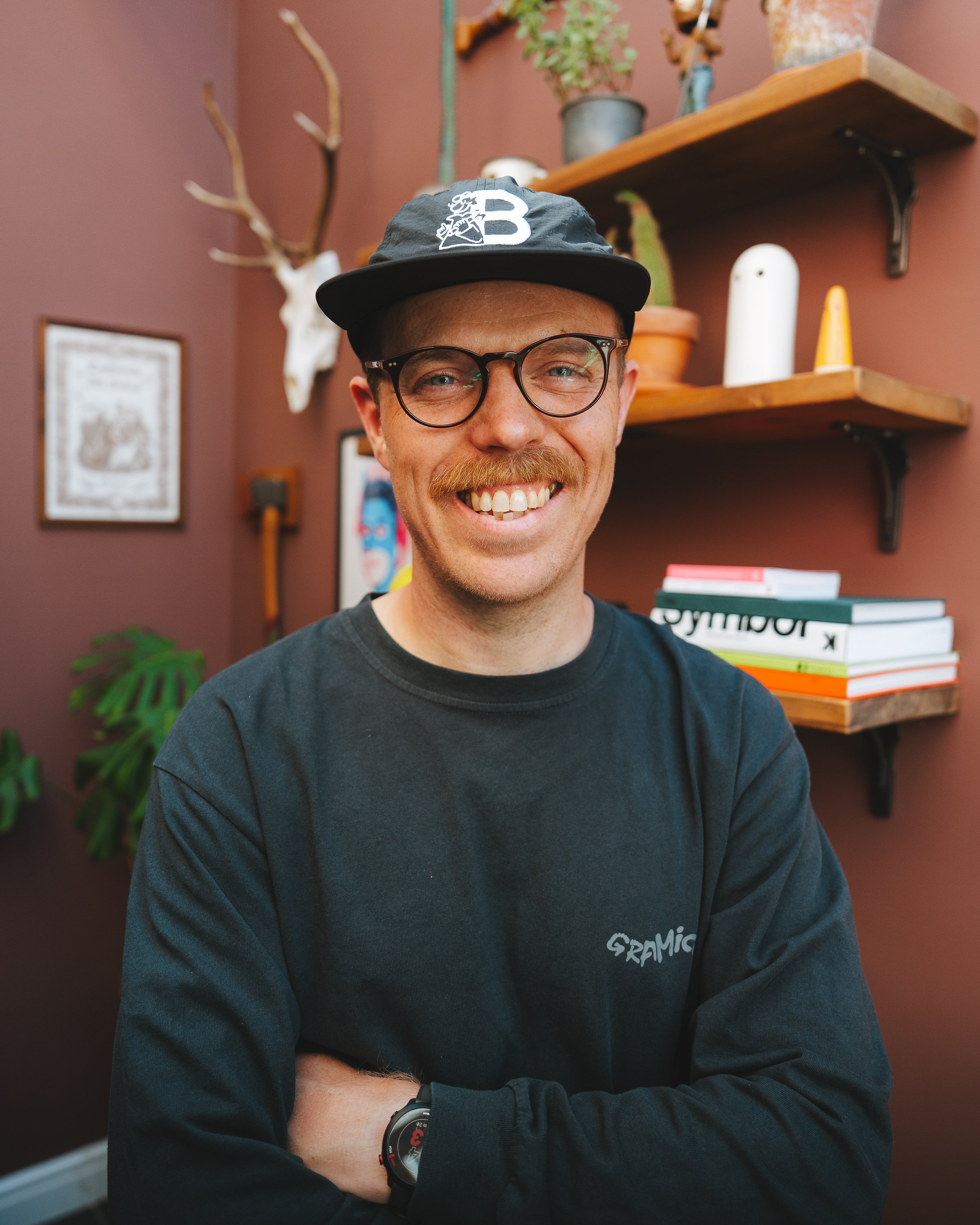 A smiling man with glasses and a beard wearing a dark baseball cap and a black sweatshirt with white text. He is standing in a room with a pinkish-brown wall, wooden shelves with potted plants, decorative items, and books.