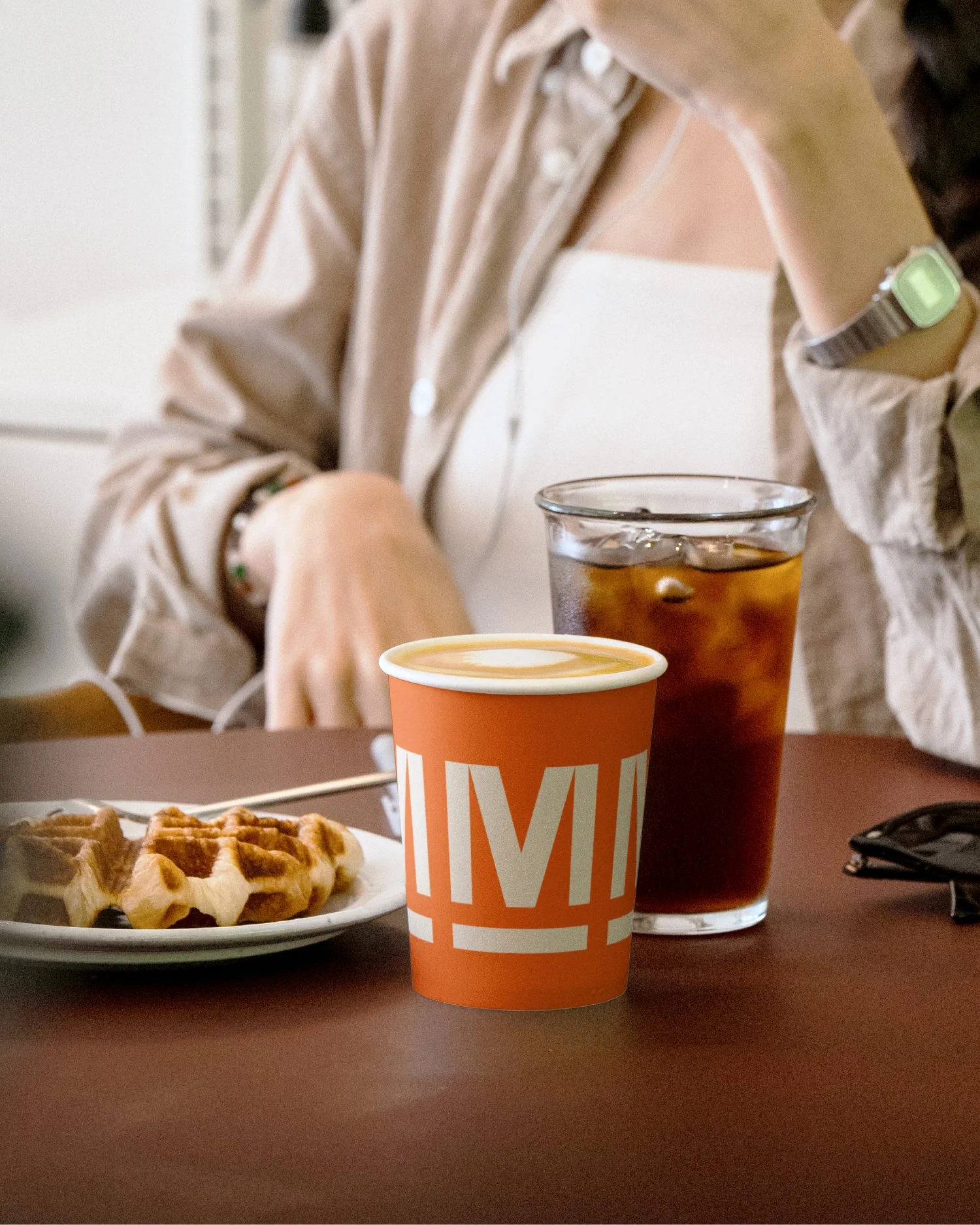 A waffle with syrup on a white plate, a cup with a warm drink, and a glass of iced soda on a brown table. A person wearing a beige shirt, white top, and a green wristwatch is seated at the table.