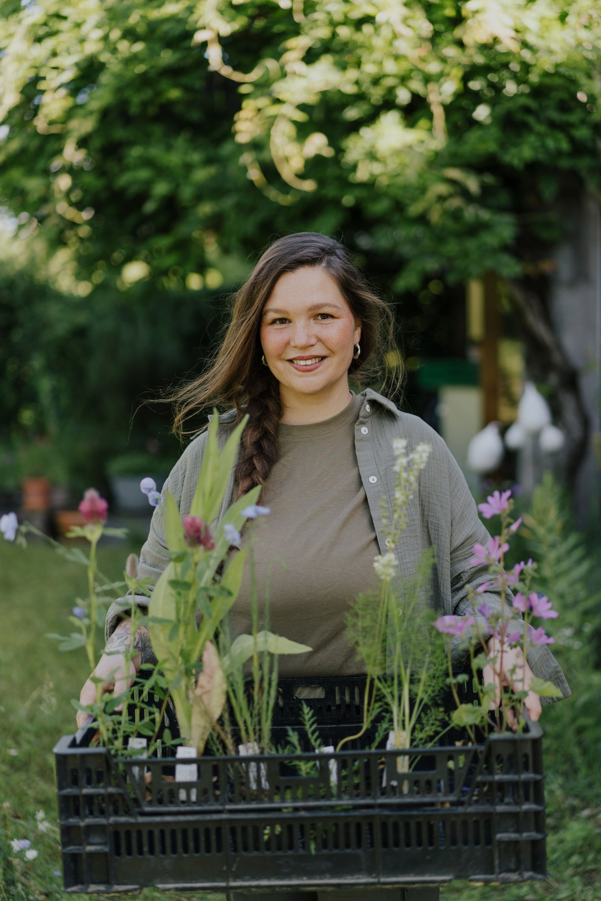 Junge Frau steht im Garten mit einen Kiste voller Wildpflanzen und Stauden, umgeben von grünen Bäumen und Pflanzen.