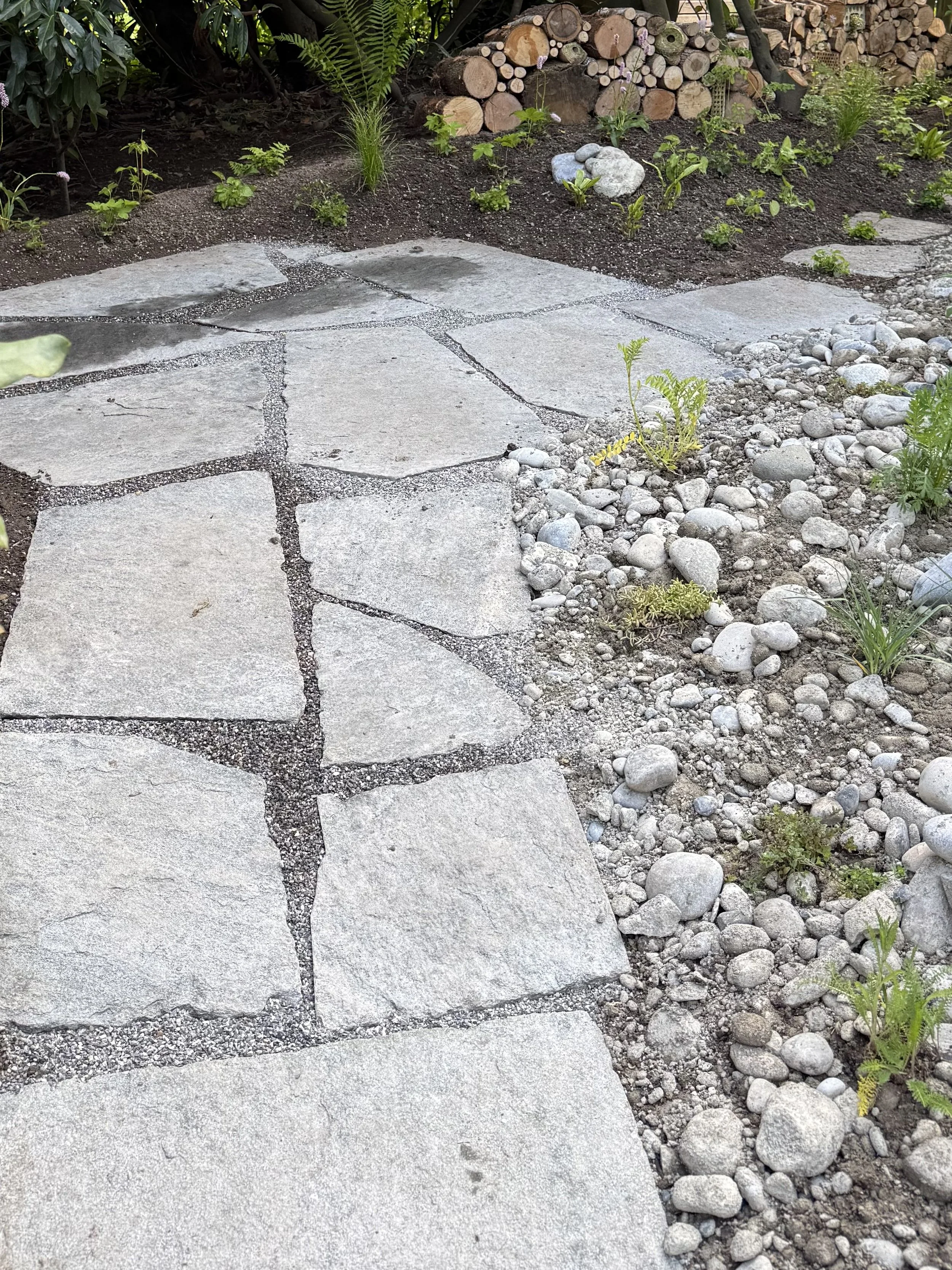 Ein Gartenweg mit Sitzplatz aus Natursteinplatten, umgeben von Erde, kleinen Steinen und Pflanzen.