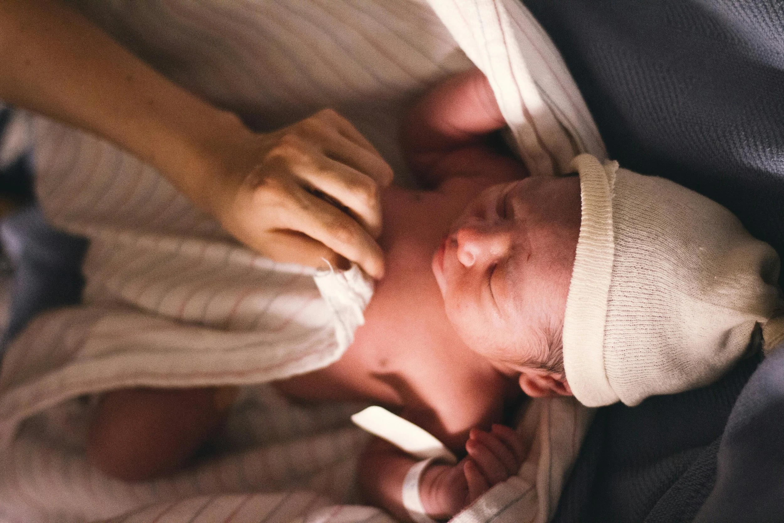 Newborn baby wrapped in a blanket wearing a soft hat, with a hand gently touching their chest.