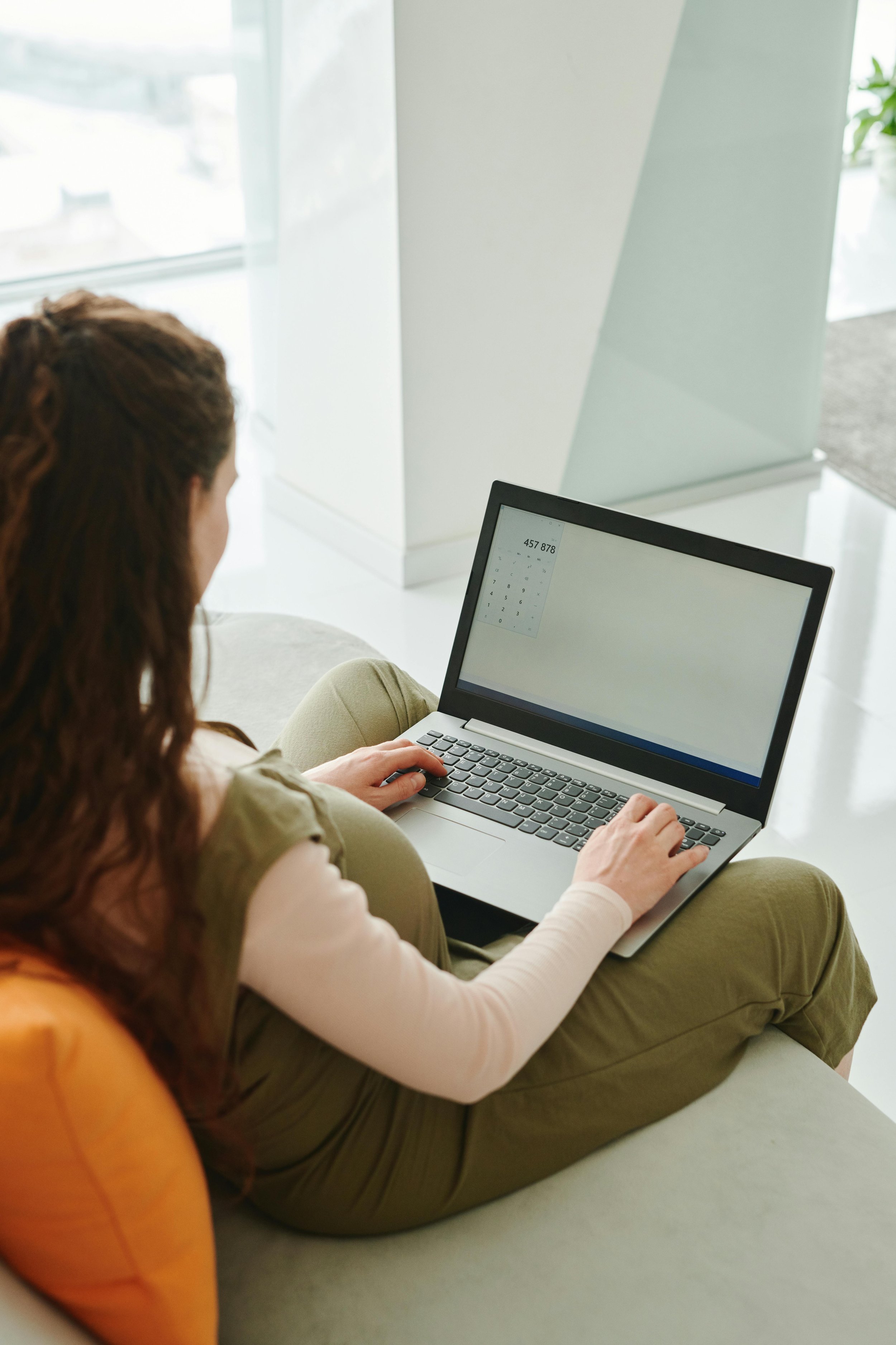 Person using a laptop with a calculator app open, sitting on a couch with an orange pillow.