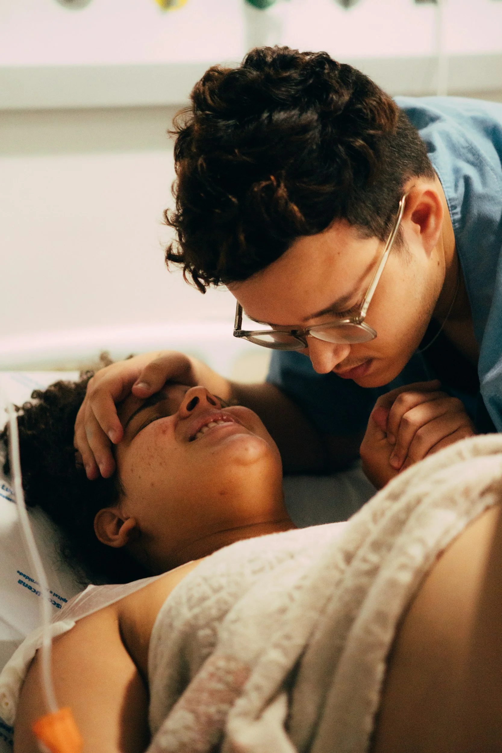 A person with glasses lying in a hospital bed, smiling and holding hands with a person who is leaning over them, possibly in a hospital or medical setting.