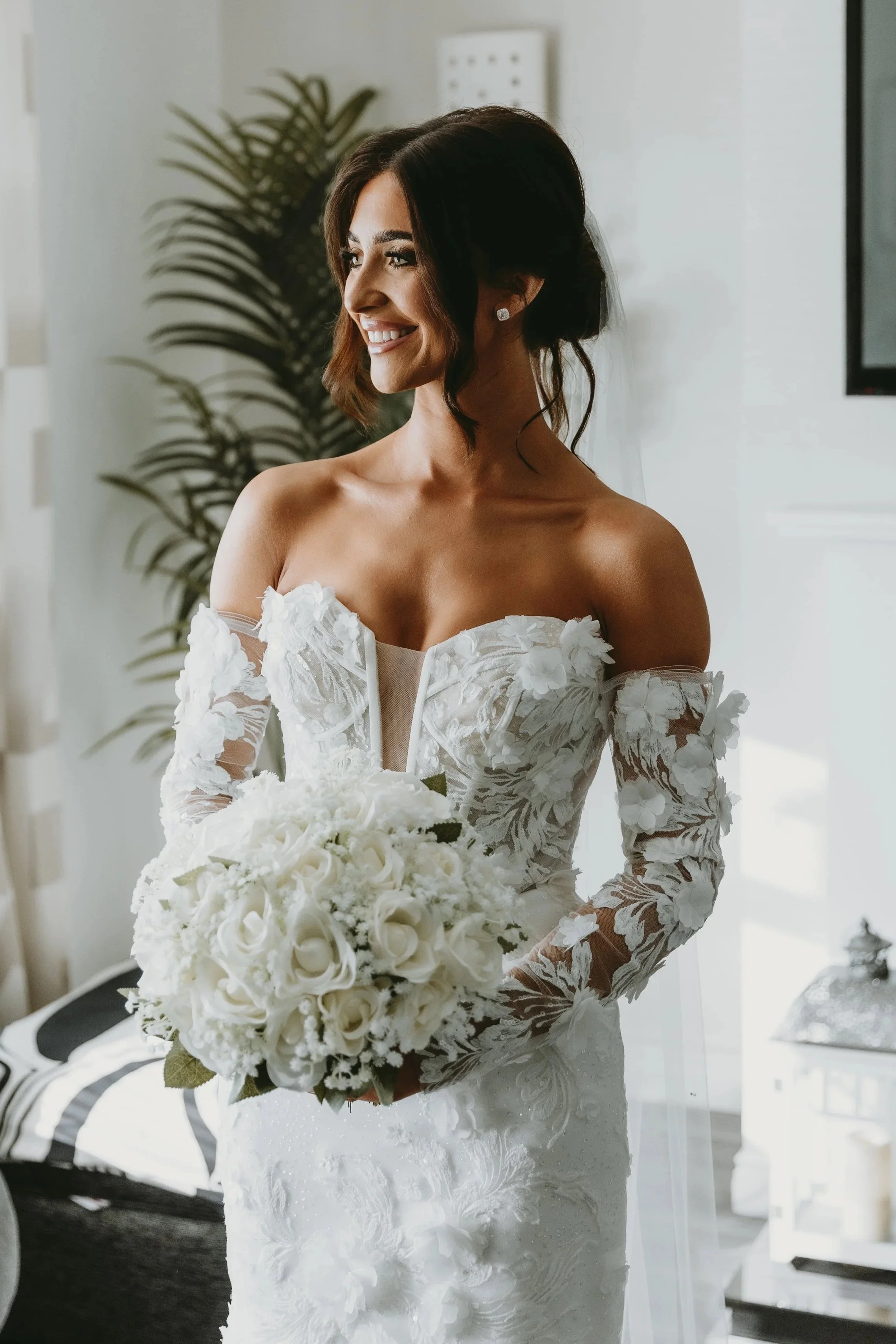 Bride in a white lace wedding dress holding a bouquet of white roses, smiling indoors with houseplants in the background.