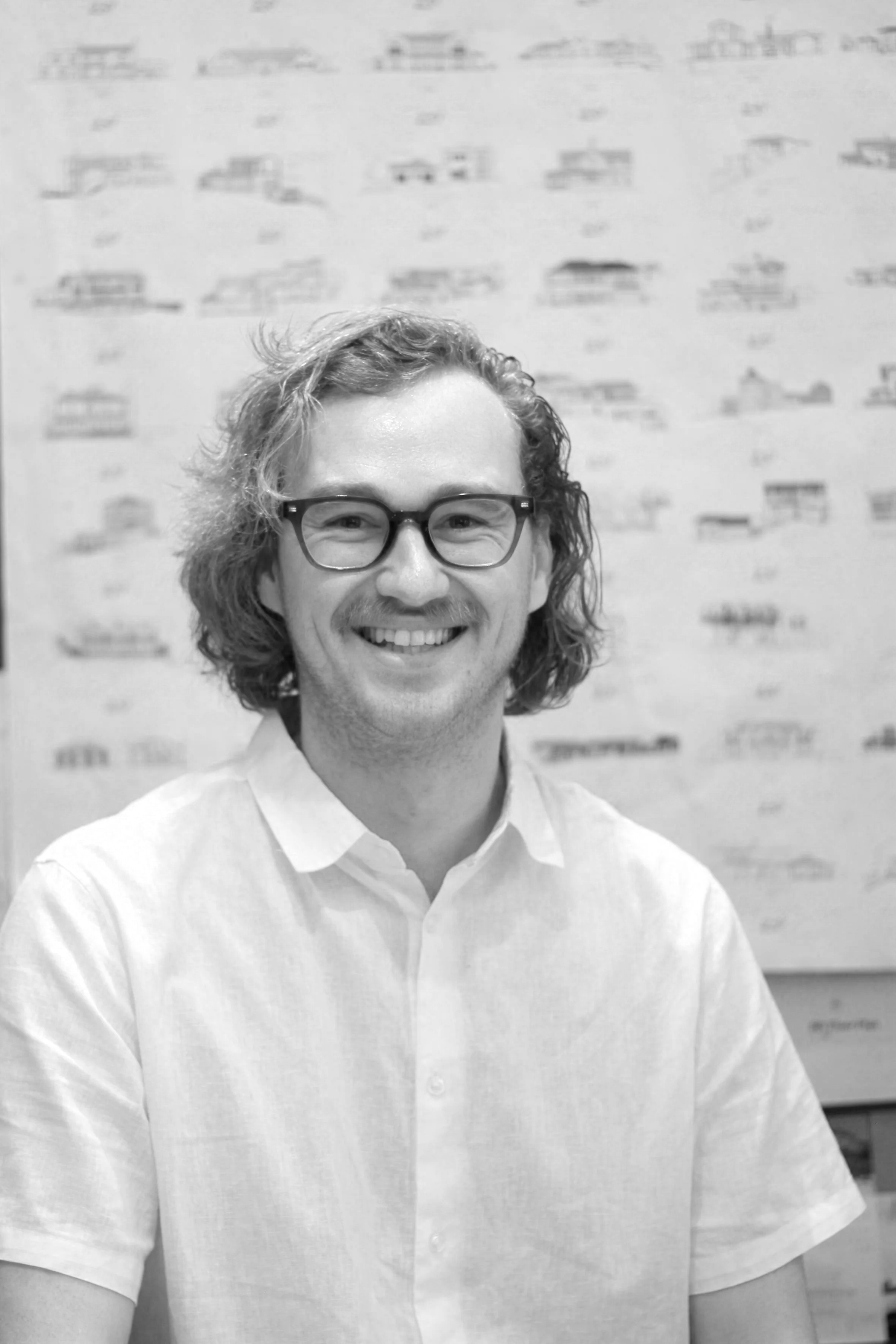 A smiling man with glasses and long hair tied back, wearing a button-up shirt, sitting in front of architectural plans or drawings.
