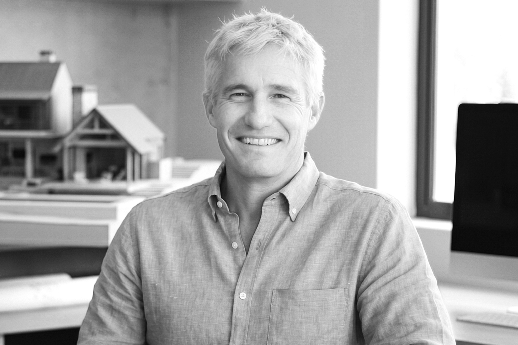 Black and white photo of a smiling man with light-colored hair wearing a button-down shirt, standing in front of a wall with sketches and printed images.