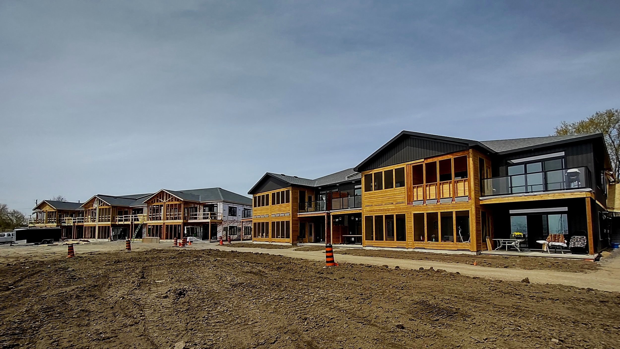 Multiple new townhouses under construction with wooden exteriors, large glass windows, and partially finished balconies, on a dirt construction site with traffic cones.