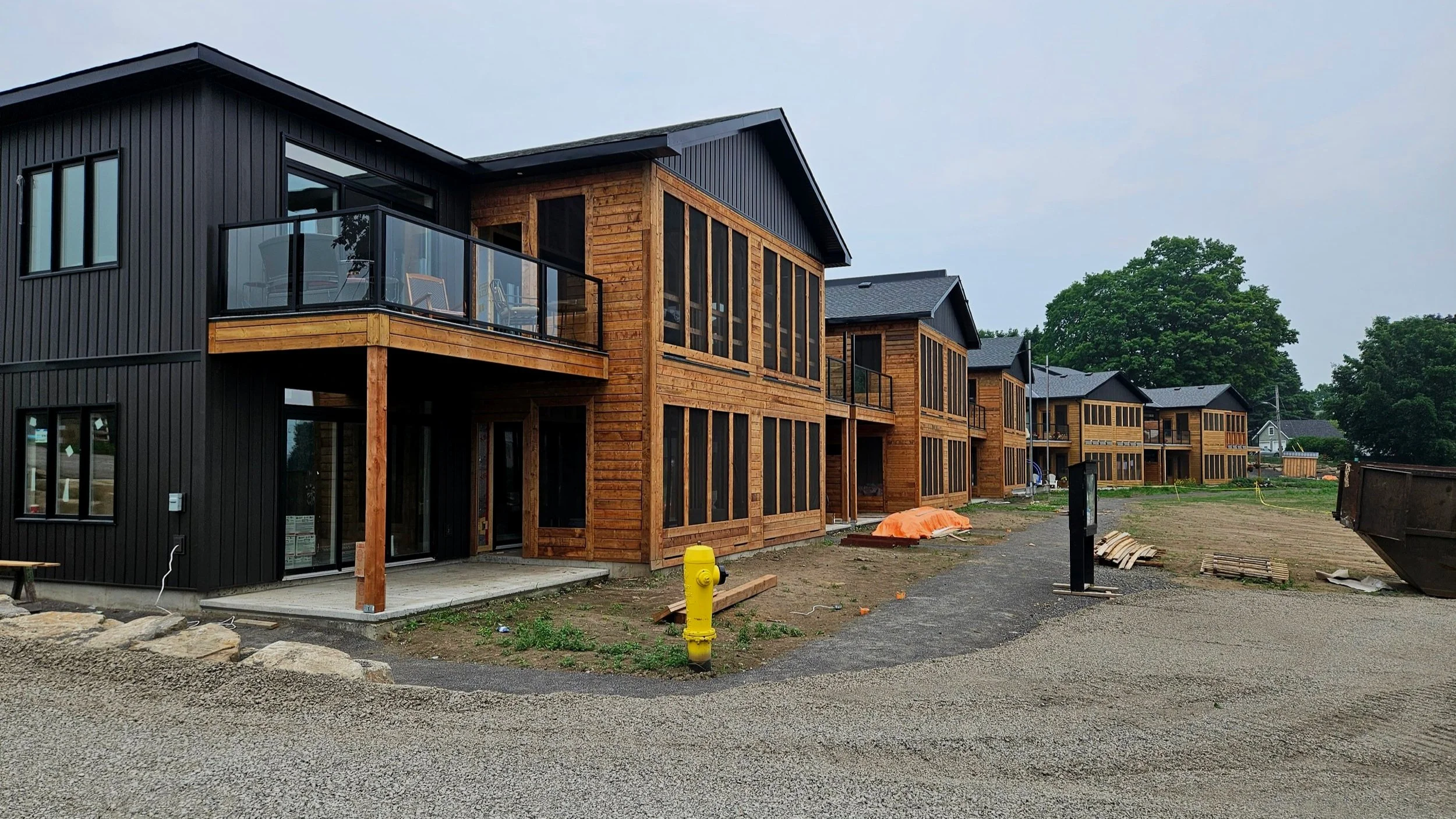 Newly constructed modern townhouses with black and wood exterior, some with balconies, on a dirt lot with construction materials and equipment, and trees in the background.