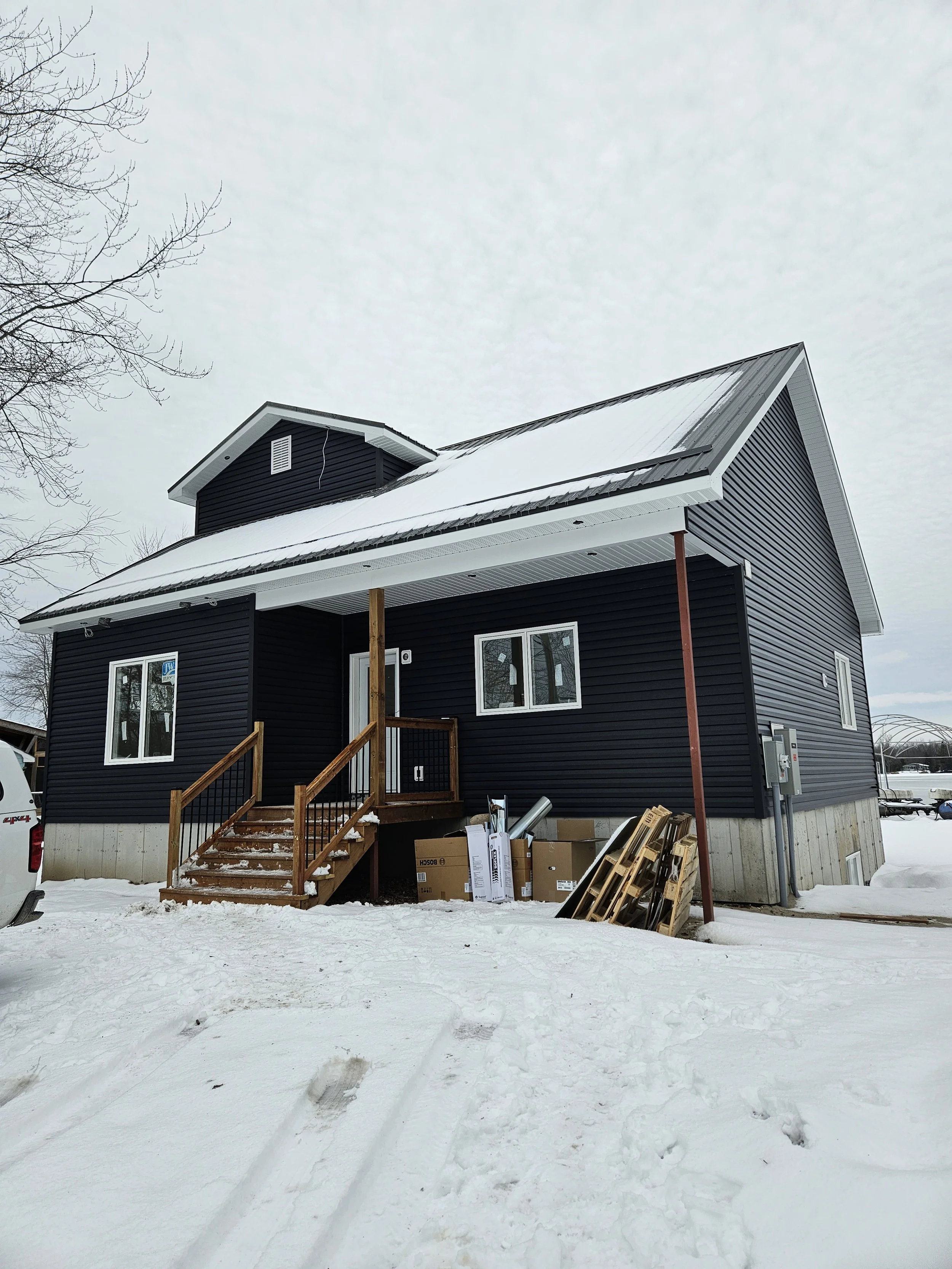 Newly constructed two-story house with black siding, white trim, metal roof, small front porch with wooden stairs, and construction materials in front, set in snowy landscape on overcast day.