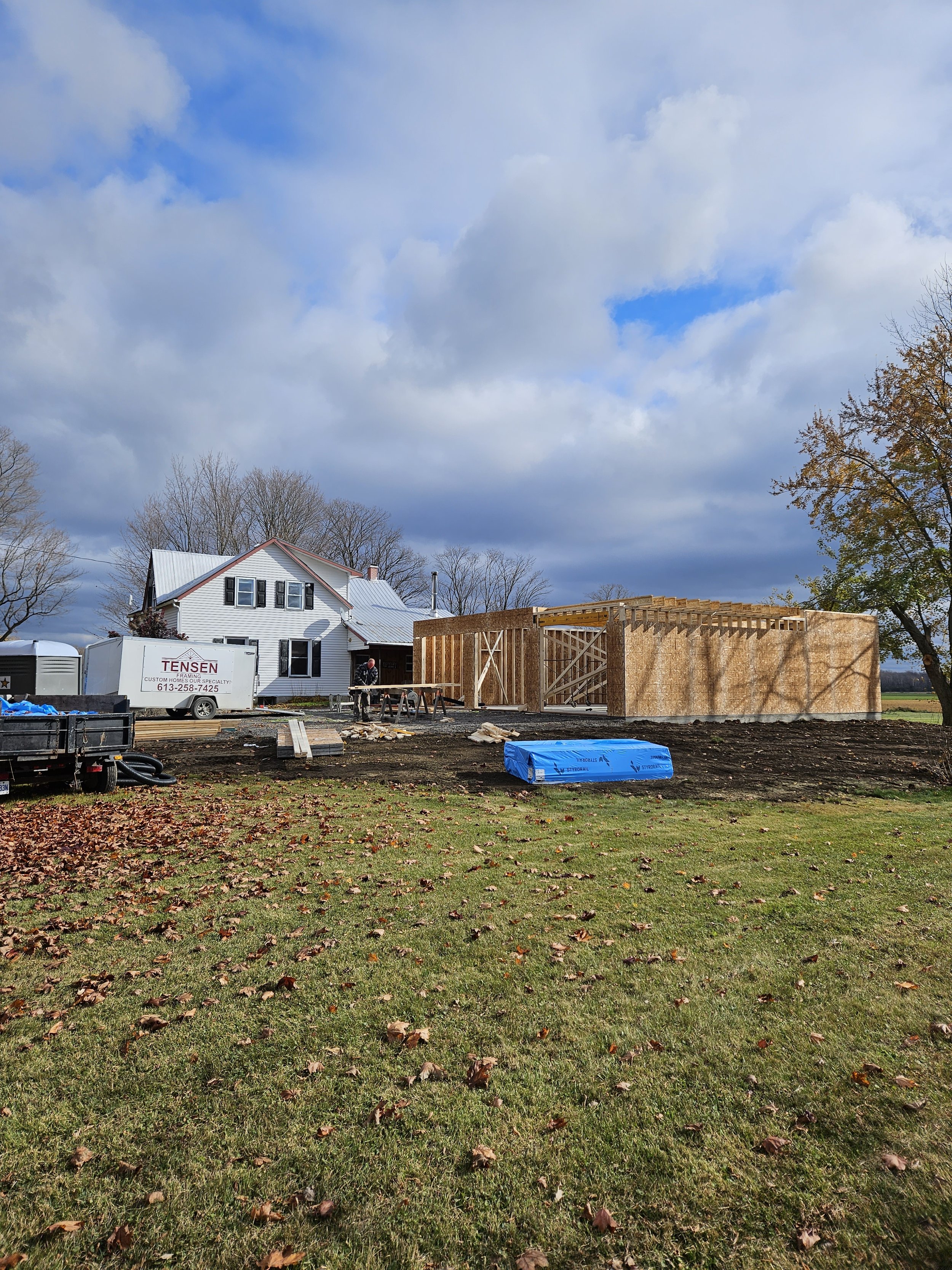 A house under construction with wooden framing, construction workers, and building equipment on a grassy yard with fallen leaves, trees, and a cloudy sky.