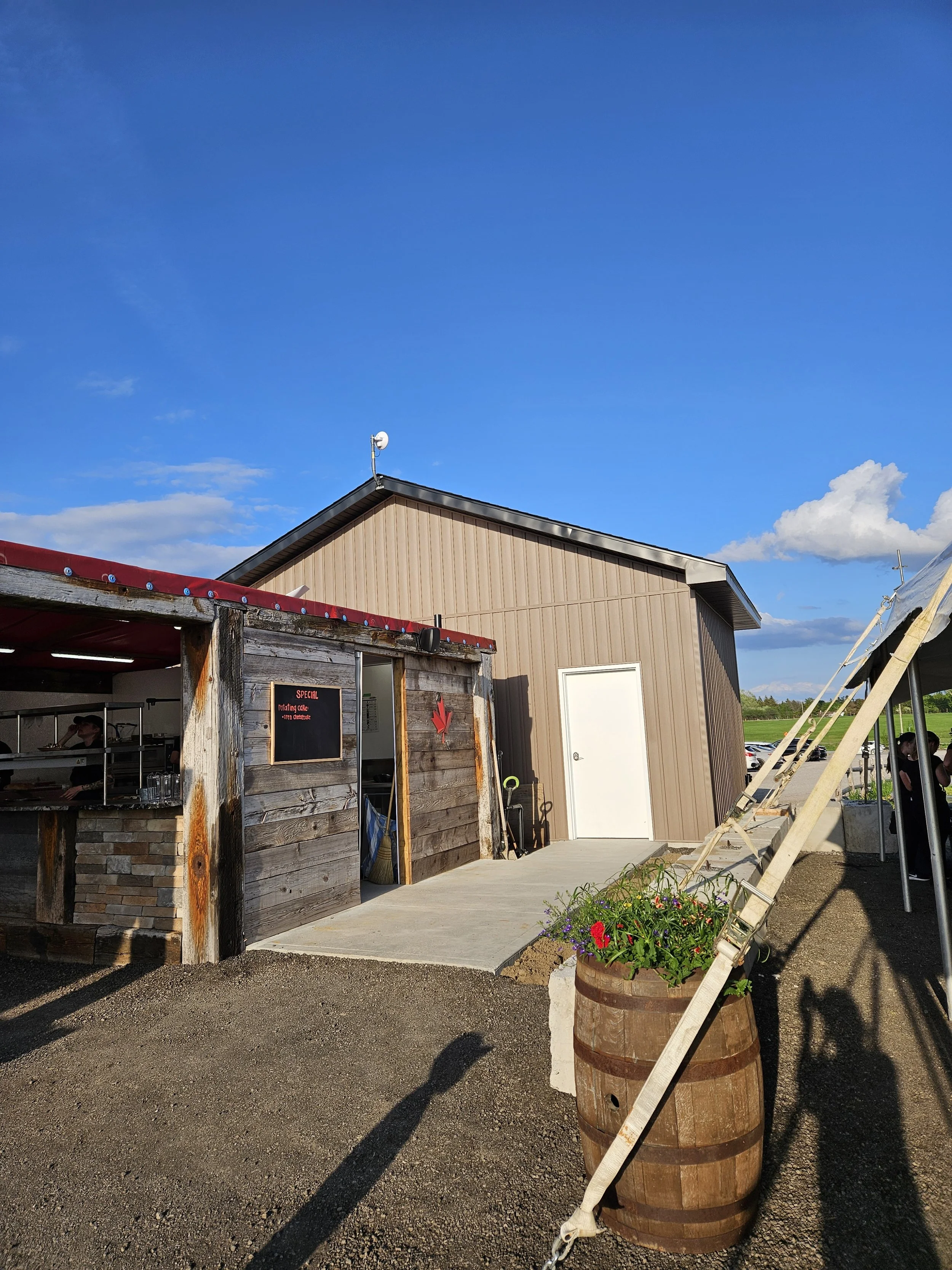 A rustic outdoor food stand with a weathered wood exterior and a black sign, along with a modern beige building and a tent on the right. There is a flower barrel planter near the walkway and clear blue sky overhead.